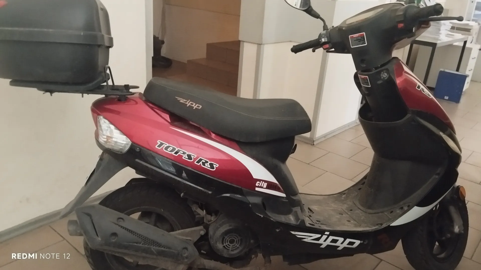 Czerwono-czarny skuter marki Zipp model TOPS RS z białymi pasami, z czarnym bagażnikiem na tylnej części. Na siedzeniu widoczny napis Zipp, na boku skutera napisy TOPS RS oraz city. Skuter stoi w pomieszczeniu z kafelkową podłogą, w tle schody i meble biurowe. W lewym dolnym rogu obrazu widoczny napis REDMI NOTE 12.