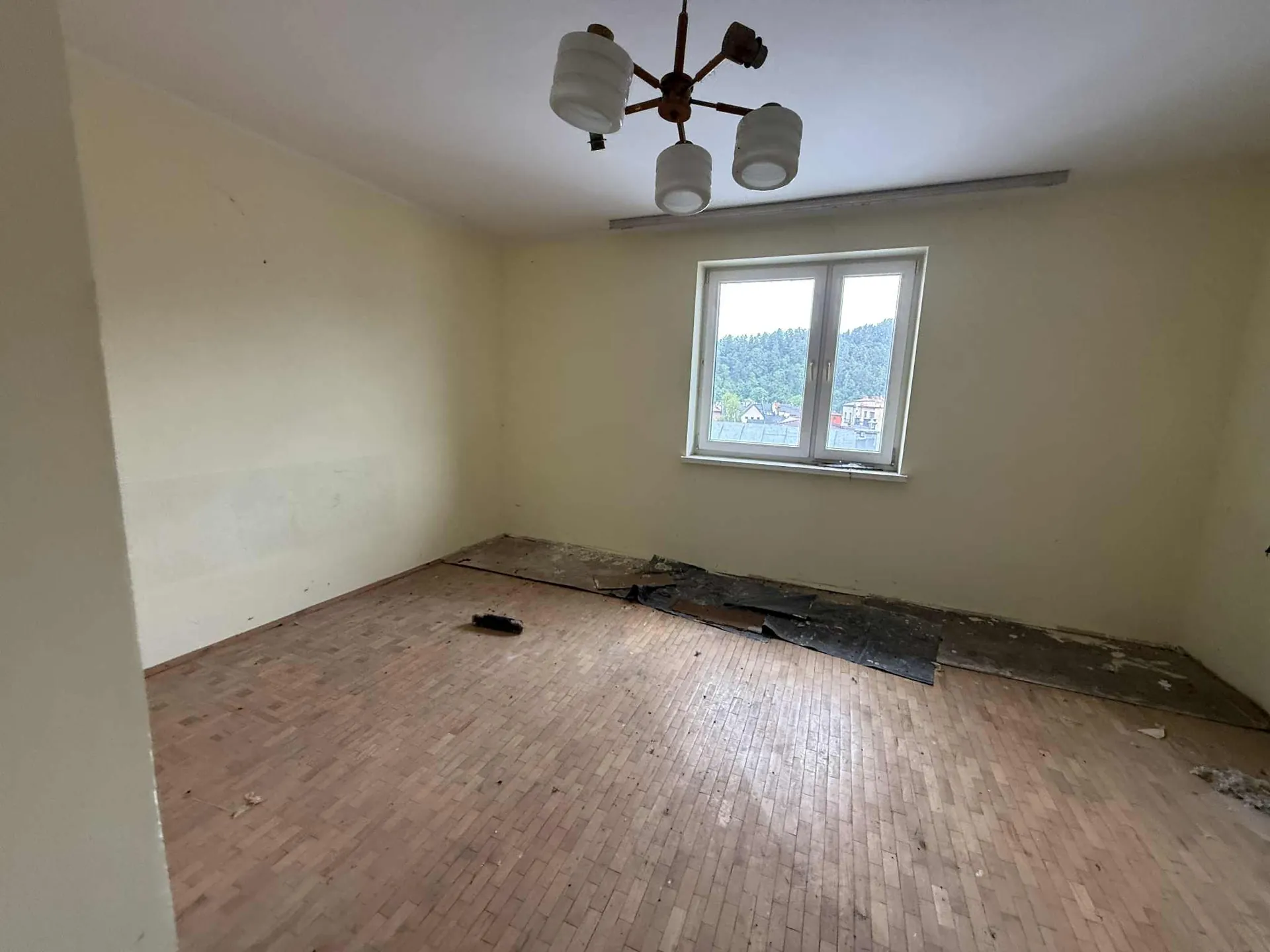 Pusty pokój z jasnożółtymi ścianami i podłogą z drewnianego parkietu. Na suficie wisząca lampa z czterema białymi kloszami. W prawej ścianie okno z widokiem na zielone wzgórza i budynki. Na podłodze w pobliżu okna uszkodzone fragmenty pokrycia oraz rozrzucone odłamki materiału. Na środku podłogi mały ciemny przedmiot.
