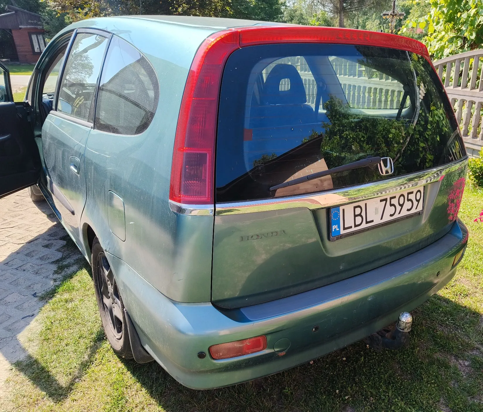 Tylny widok zielonego samochodu typu kombi zaparkowanego na trawie obok kostki brukowej. Samochód ma otwarte przednie lewe drzwi. Na klapie bagażnika widnieje napis HONDA oraz polska tablica rejestracyjna o numerze LBL 75959. Pojazd posiada hak holowniczy.