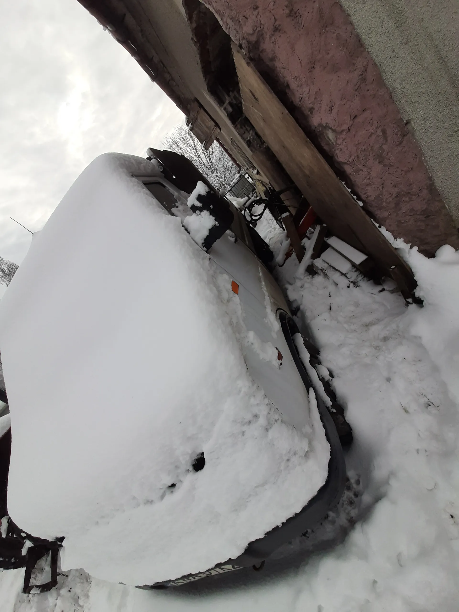 Biały samochód całkowicie pokryty śniegiem, stojący obok budynku z różową ścianą i drewnianymi deskami. W tle widoczne są nagie drzewa i pochmurne niebo. Śnieg pokrywa również ziemię i inne przedmioty w otoczeniu.
