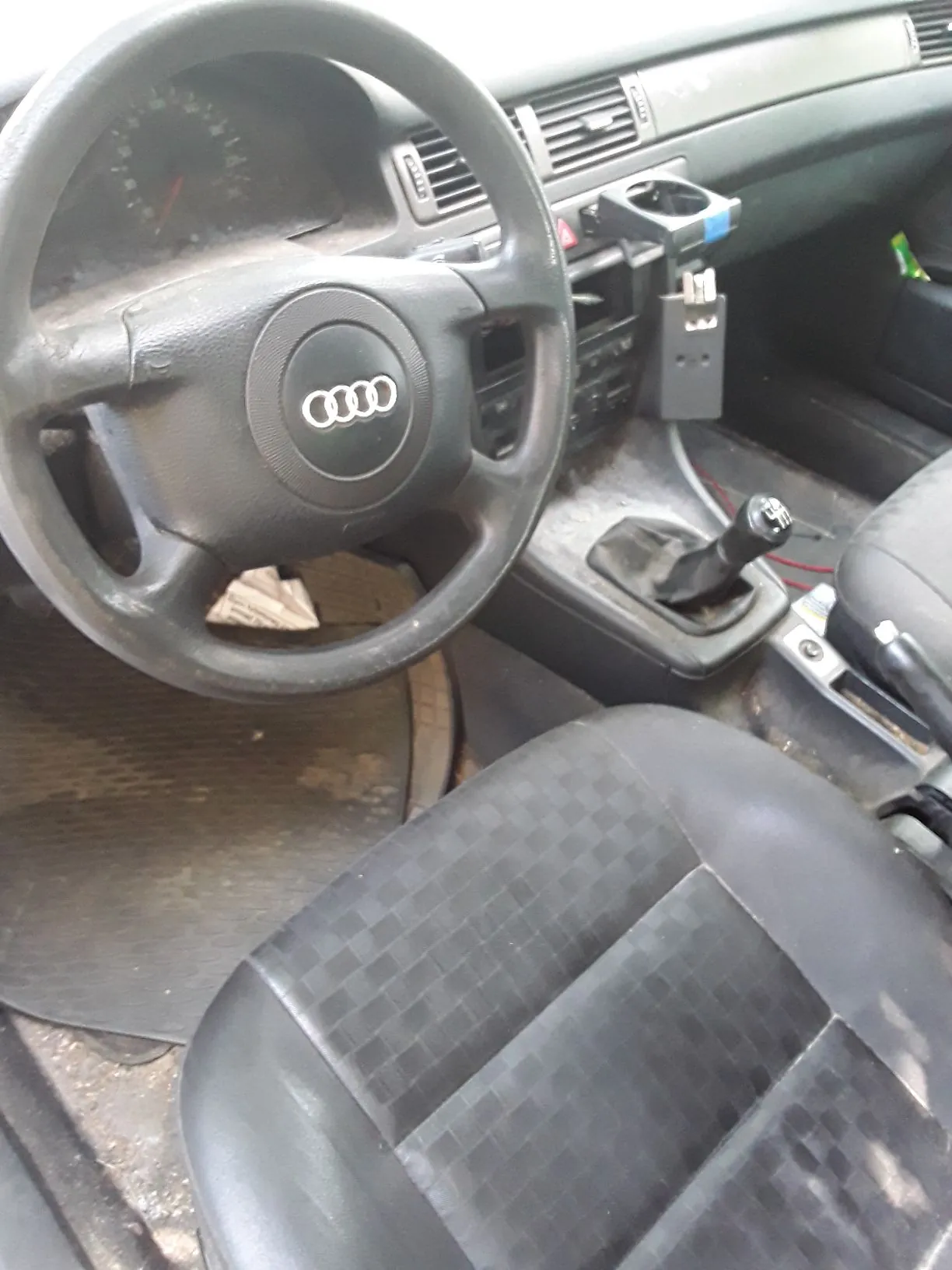 Wnętrze samochodu marki Audi z perspektywy kierowcy. Widoczna jest czarna kierownica z logo Audi w centrum, deska rozdzielcza, lewarek zmiany biegów oraz czarny, skórzany fotel z przodu. W uchwycie na kubki znajduje się czarny przedmiot.