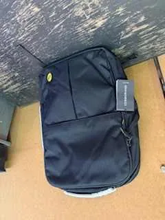 Czarny plecak z przodem kieszenią na suwak, żółtym okrągłym logo i szarą etykietą z napisem 'LAPTOP BAG'. Plecak leży na powierzchni z drewnianym wzorem i brązowym tłem.