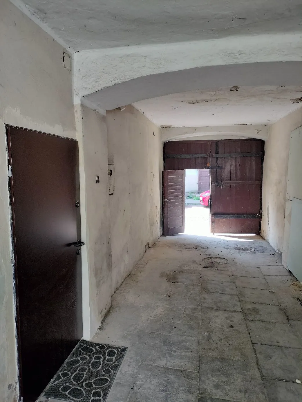Wnętrze jasnego, surowego korytarza z tynkowanymi ścianami i betonową podłogą. Po lewej stronie znajduje się ciemnobrązowe drzwi, a przed nimi leży szara wycieraczka z białym wzorem. Na końcu korytarza widoczne są duże, drewniane, brązowe wrota, z których mniejsza część jest otwarta, ukazując jasne światło dzienne i fragment czerwonego przedmiotu na zewnątrz.