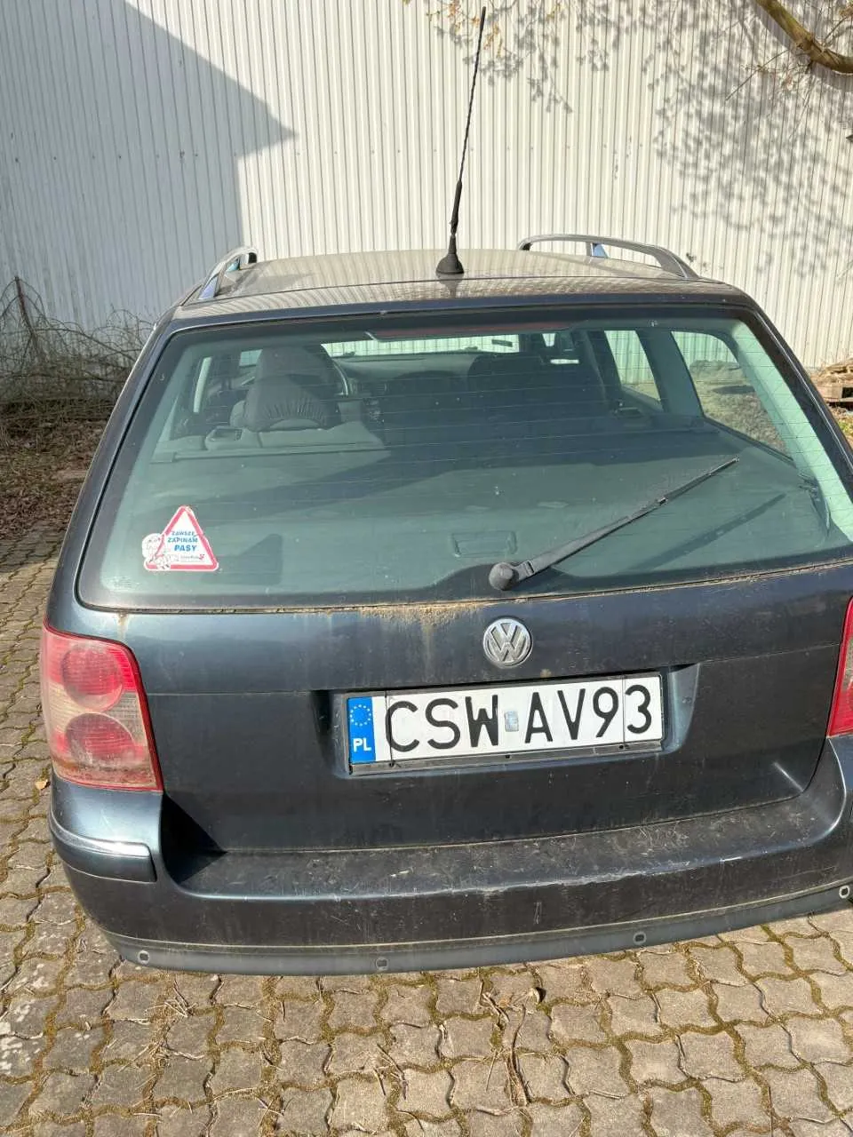 Tył ciemnoszarego samochodu osobowego marki Volkswagen z tablicą rejestracyjną CSW AV93 z polskim oznaczeniem PL. Na tylnej szybie widoczny czerwony trójkątny naklejka z napisem. Samochód posiada dachowy bagażnik i antenę. Tło stanowi biała metalowa ściana i brukowana powierzchnia. Wnętrze samochodu częściowo widoczne przez szybę.