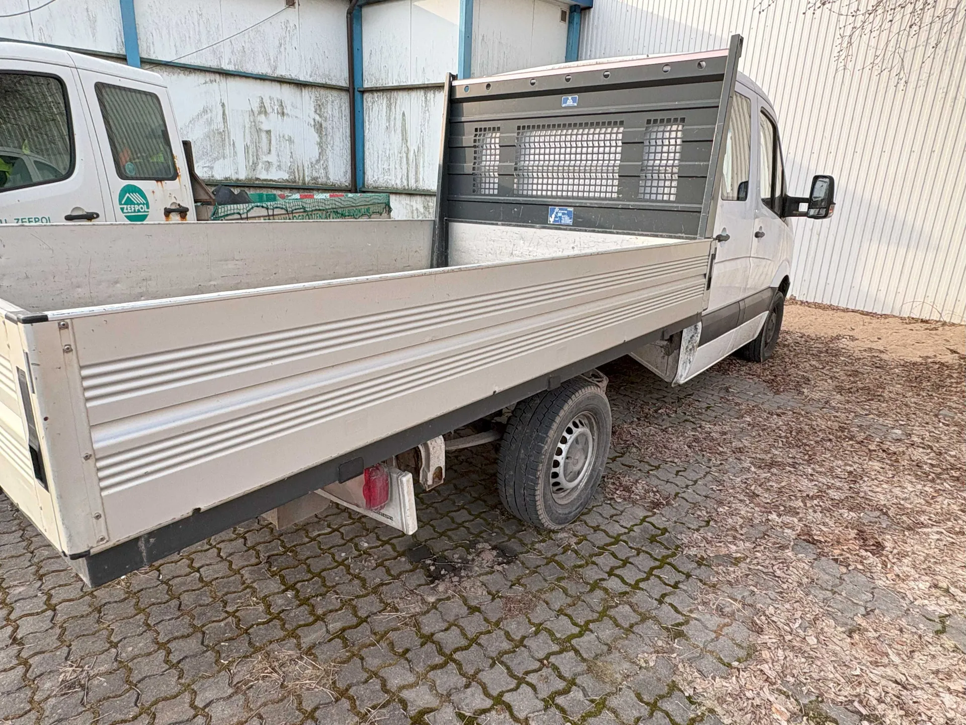 Biały samochód dostawczy z otwartym, pustym ladunkiem o metalowych burtach z poziomymi żebrami. Widoczne są czerwone światła tylnego oraz miejsce na tablicę rejestracyjną. W tle budynek z białymi i niebieskimi elementami, częściowo zardzewiałymi. Po lewej stronie fragment innego białego pojazdu z zielonym logo 'ZEPOL'. Podłoga z bruku kamiennego pokryta suchymi liśćmi. Na ścianie budynku widoczna siatka i zielono-czerwony element.