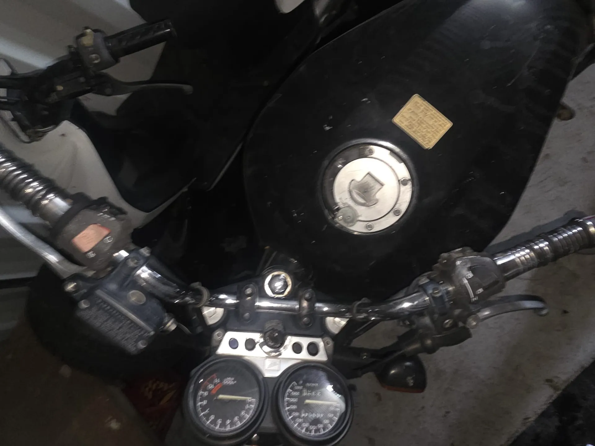 Zbliżenie na kierownicę motocykla z dwoma okrągłymi wskaźnikami prędkości i obrotomierzem po lewej stronie. Czarny zbiornik paliwa z srebrnym korkiem i żółtą etykietą. Metalowe kierownice z przyciskami, dźwigniami oraz czarnymi uchwytami. W tle widoczna betonowa podłoga.