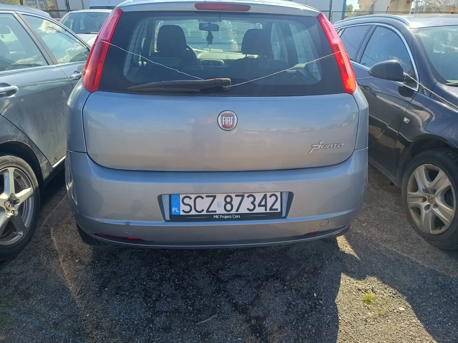 Tylny widok srebrnego samochodu marki Fiat Punto zaparkowanego między dwoma innymi pojazdami. Na klapie bagażnika znajduje się logo FIAT oraz napis PUNTO. Samochód posiada polską tablicę rejestracyjną o numerze SCZ 87342, a pod nią widnieje napis MK Project Cars.