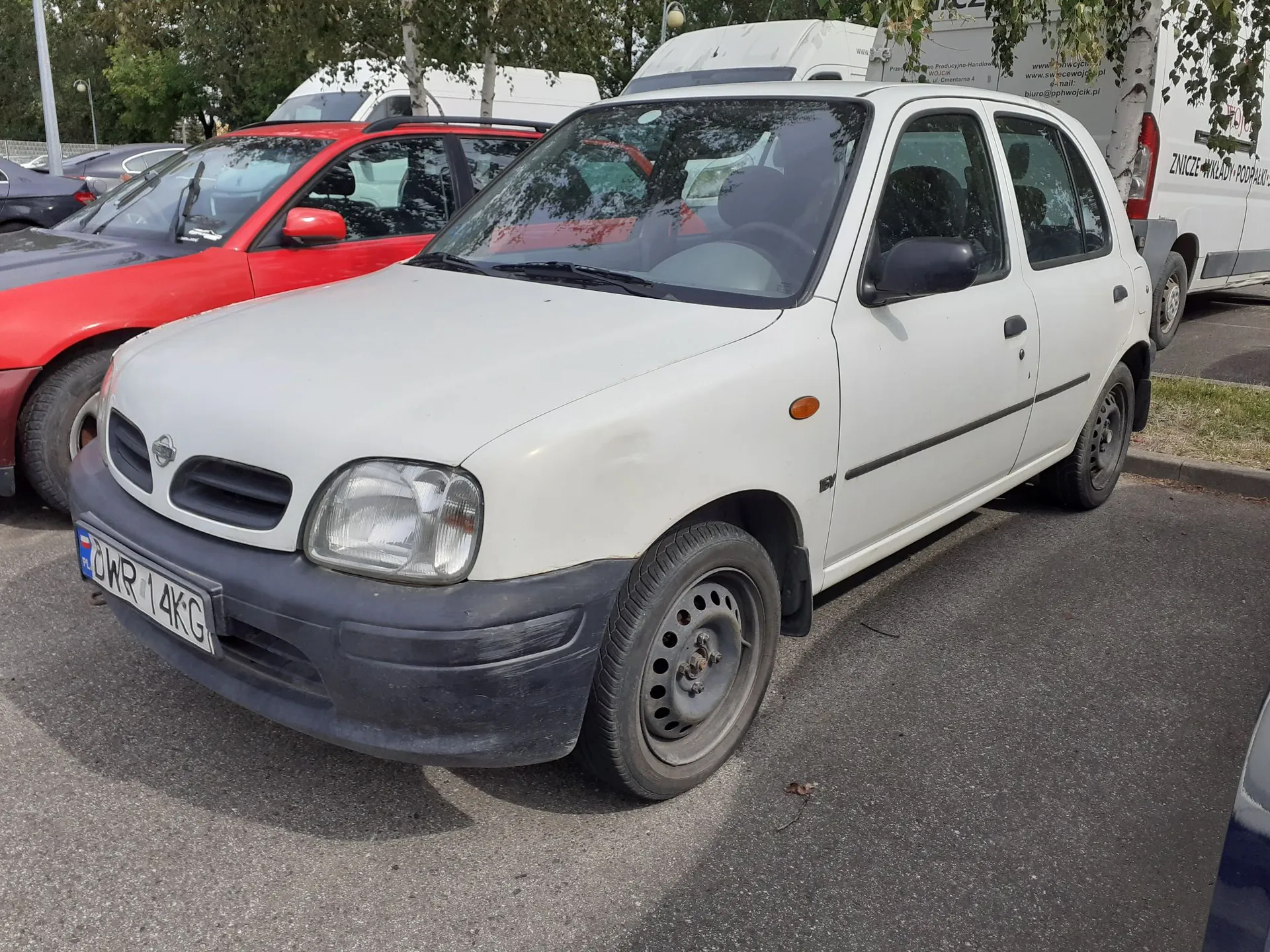 Biały, niewielki samochód osobowy zaparkowany na asfaltowym parkingu. Pojazd ma czarny zderzak przedni oraz czarne listwy boczne. Widoczna jest tablica rejestracyjna o numerze CWR 14KG. Obok białego auta stoi czerwony samochód osobowy, a w tle widoczne są białe furgonetki oraz drzewa.