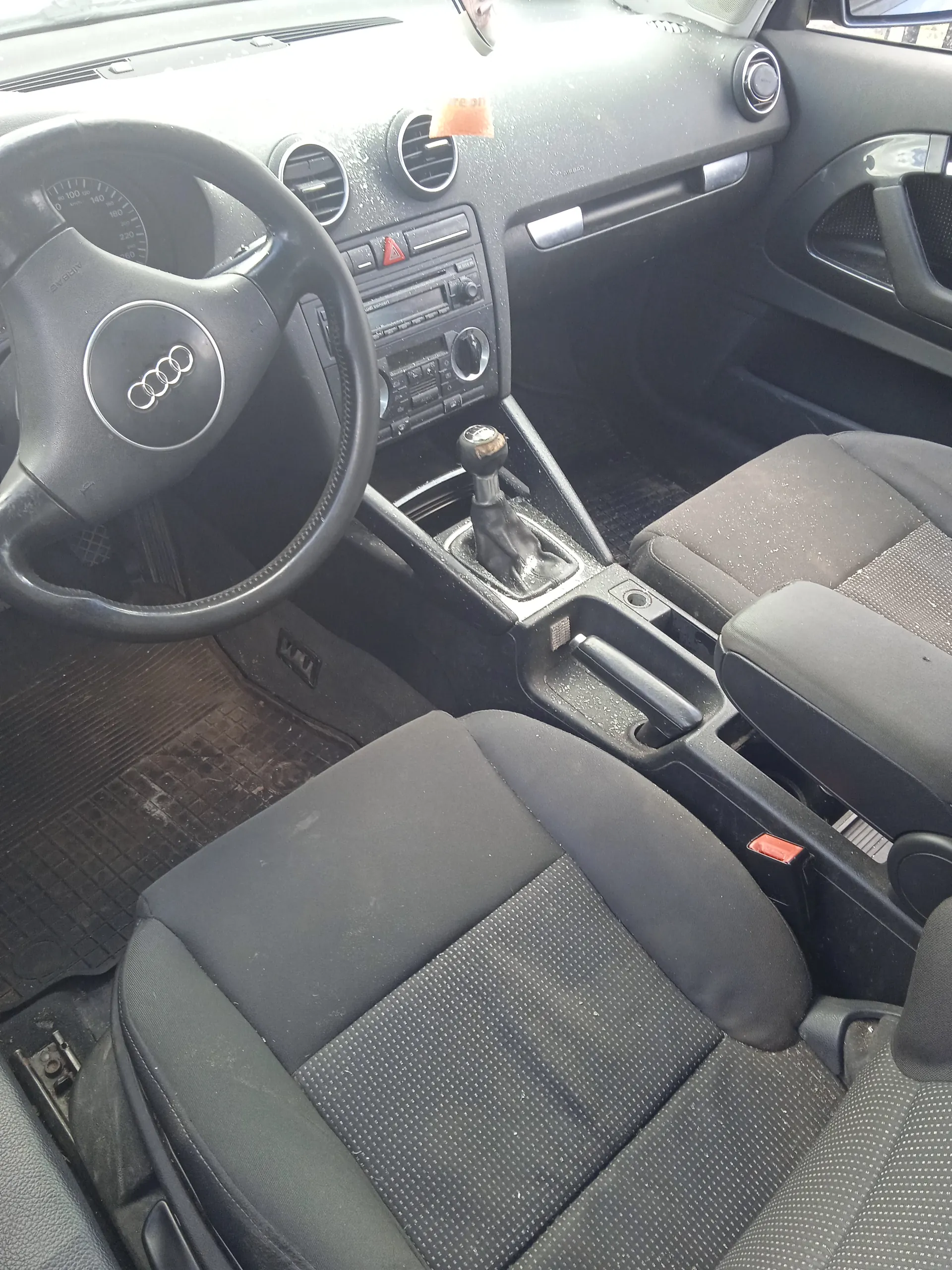 Wnętrze samochodu marki Audi z widocznym kierownicą z logo czterech pierścieni, czarnymi siedzeniami z tkaniny w drobny wzór, manualną skrzynią biegów, deska rozdzielczą z prędkościomierzem, wentylacjami i radiem. Podłoga pokryta brudnymi matach. Wnętrze wydaje się nieco zabrudzone.