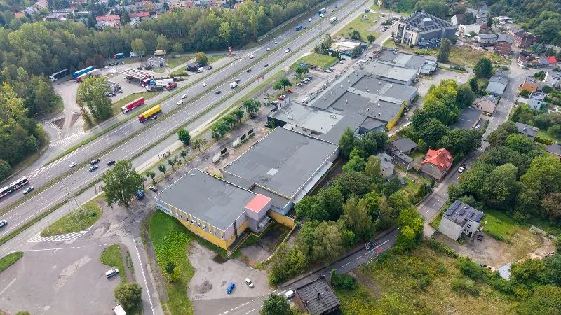 Widok z lotu ptaka na obszar przemysłowo-handlowy przy autostradzie. Po lewej stronie przebiega wielopasmowa droga z jadącymi samochodami i ciężarówkami. W centrum znajduje się kompleks dużych, płaskich budynków magazynowych i hal o szarych dachach, z których niektóre mają żółte elewacje. Wokół budynków widoczne są parkingi, place manewrowe dla ciężarówek oraz obszary zielone z drzewami. W tle oraz po prawej stronie znajdują się mniejsze budynki mieszkalne i domy jednorodzinne z czerwonymi dachami, otoczone gęstą roślinnością.