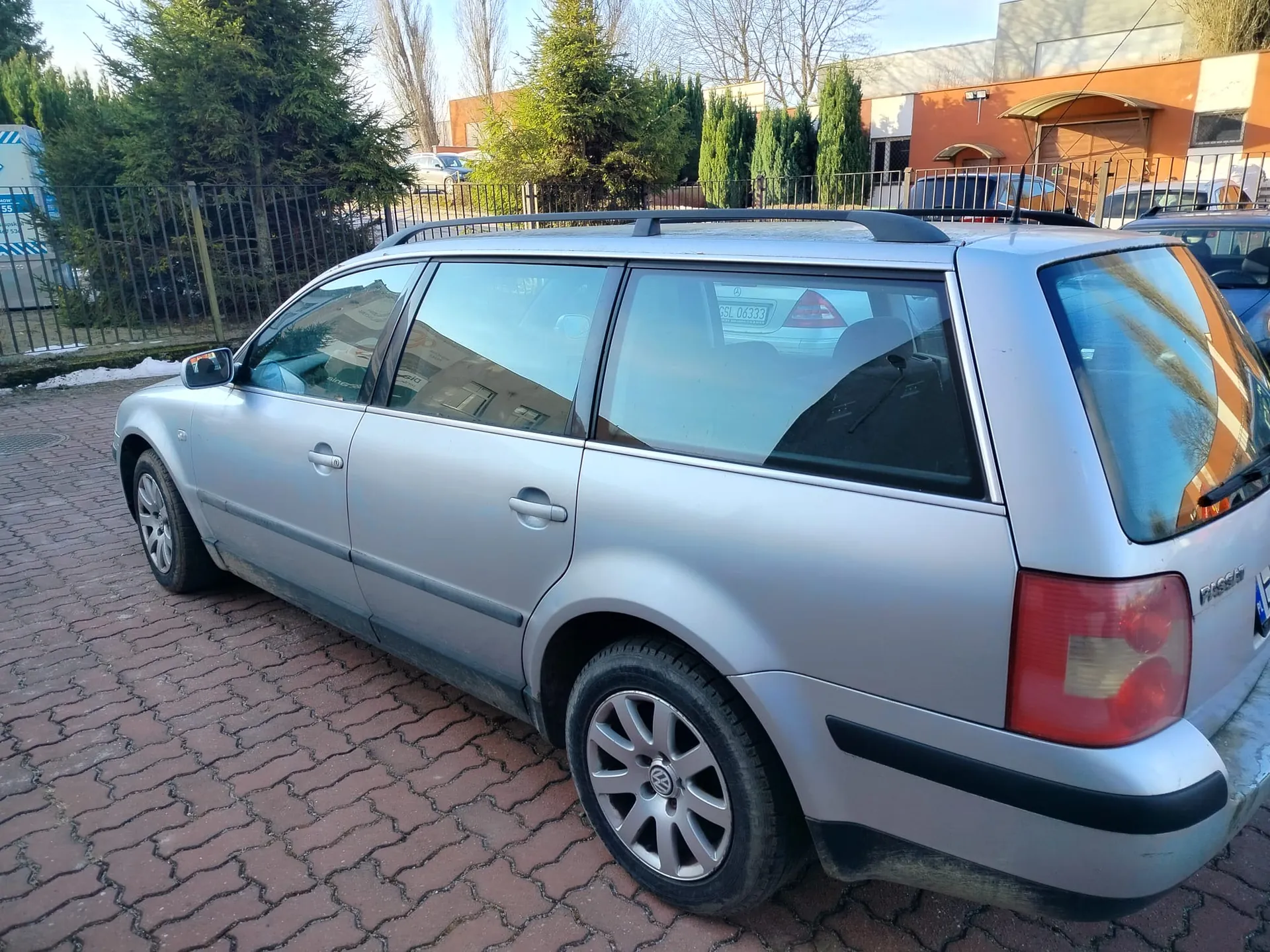 Srebrny samochód typu kombi Volkswagen Passat zaparkowany na brukowanym placu. Pojazd posiada dachówki, felgi aluminiowe oraz widoczne światła tylne. W tle metalowe ogrodzenie, iglaste drzewa, budynki o pomarańczowo-białych elewacjach oraz inne zaparkowane samochody. Na szybie tylnej odbija się tablica rejestracyjna innego pojazdu z napisem GSL 0633. Niebo jest bezchmurne.