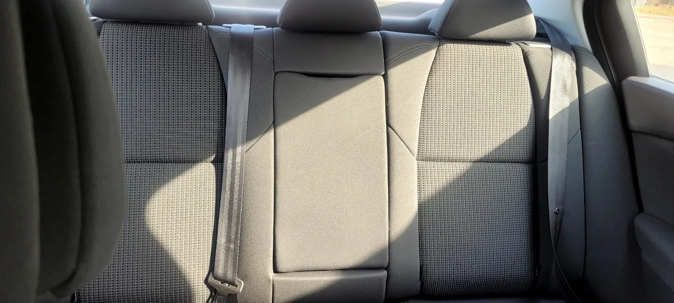 Wnętrze samochodu z widokiem na tylną kanapę. Trzy siedzenia z poduszkami pod głowę, pokryte tkaniną w drobny wzór. Środkowe siedzenie ma gładką, jednolitą powierzchnię. Widoczne pasy bezpieczeństwa po lewej i prawej stronie. Oświetlenie słoneczne rzuca cienie na siedzenia. Wnętrze w odcieniach szarości.