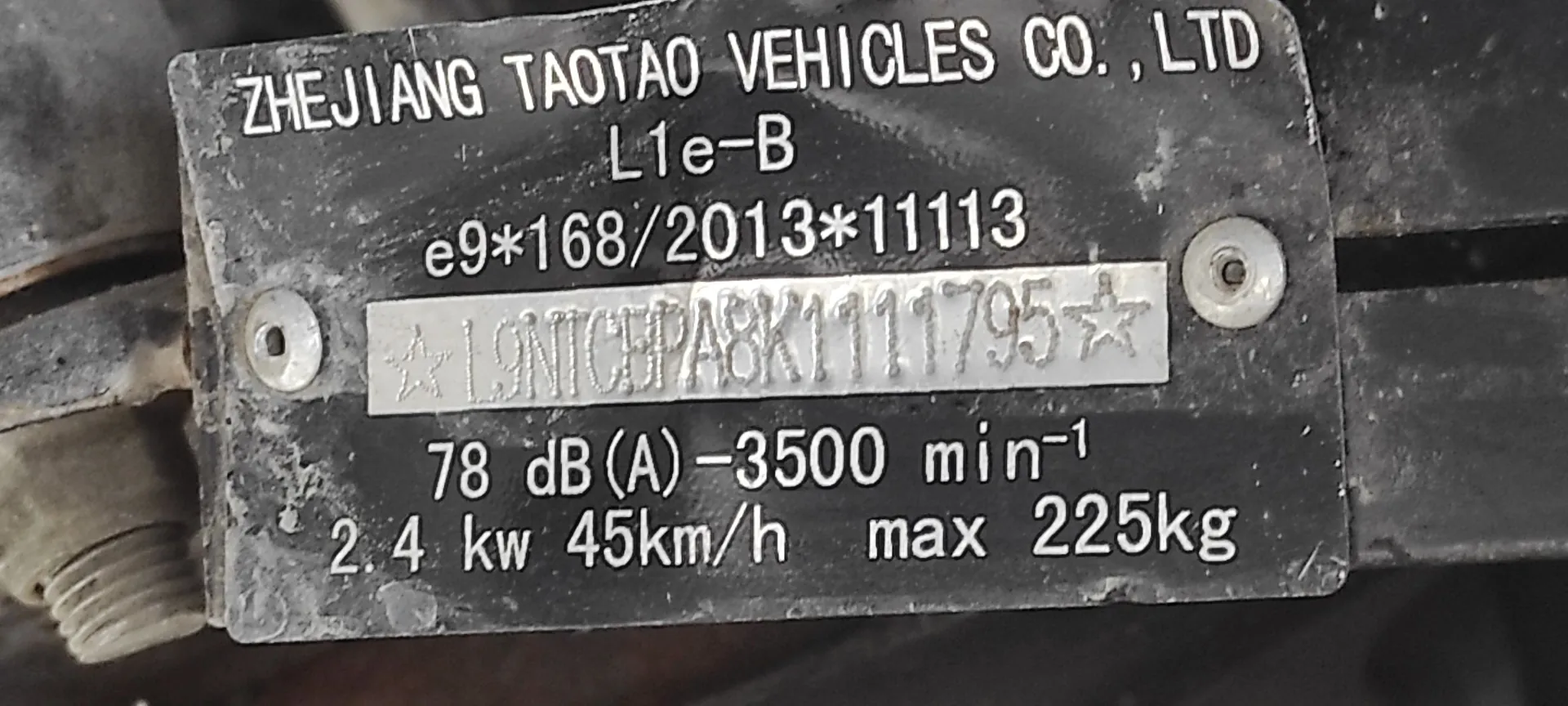 Metalowa tabliczka znamionowa pojazdu z białymi napisami na czarnym tle. Tekst na tabliczce brzmi: 'ZHEJIANG TAOTAO VEHICLES CO., LTD', poniżej 'L1e-B', następnie 'e9*168/2013*11113'. Poniżej znajduje się numer seryjny w ramce: 'L9NTCBPAS K1111795'. Na dole widnieją parametry techniczne: '78 dB (A) - 3500 min-1' oraz '2.4 kw 45km/h max 225kg'. Tabliczka jest przymocowana za pomocą dwóch śrub po bokach.