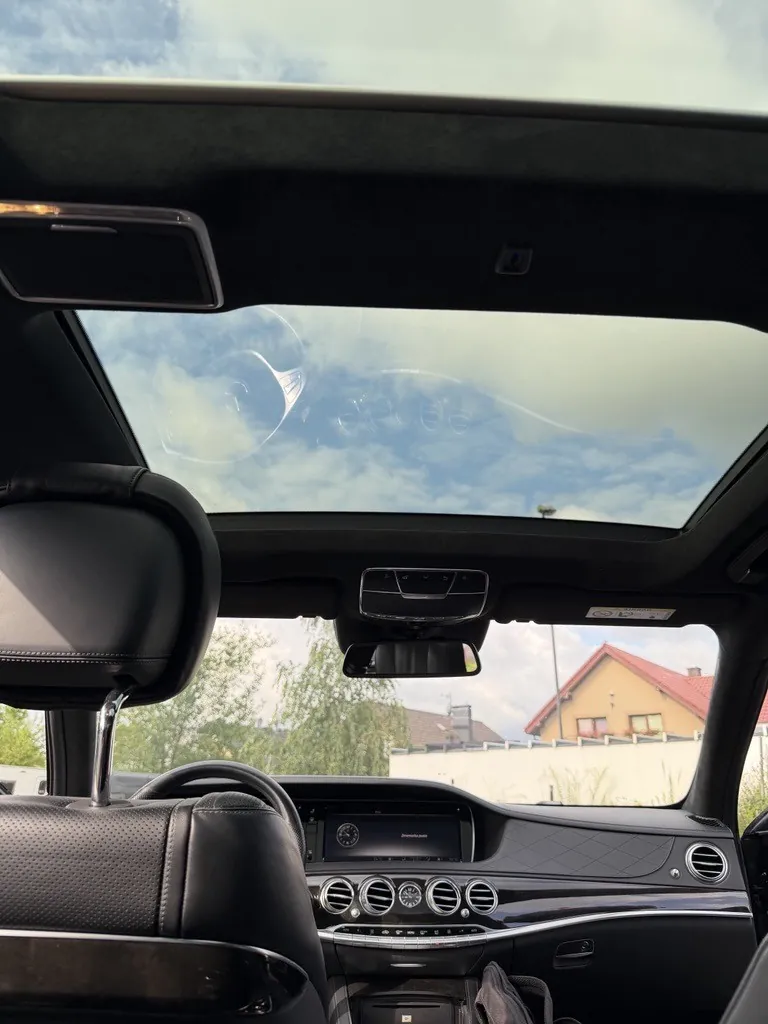 Widok z wnętrza samochodu przez panoramiczny dach. W górnej części zdjęcia widoczne jest niebo z chmurami przez szklany dach. Poniżej znajduje się czarny zagłówek fotela kierowcy, fragment kierownicy oraz deska rozdzielcza z ekranem multimedialnym i okrągłymi wylotami nawiewów. Przez przednią szybę widoczny jest dom z czerwonym dachem oraz zielone drzewa.