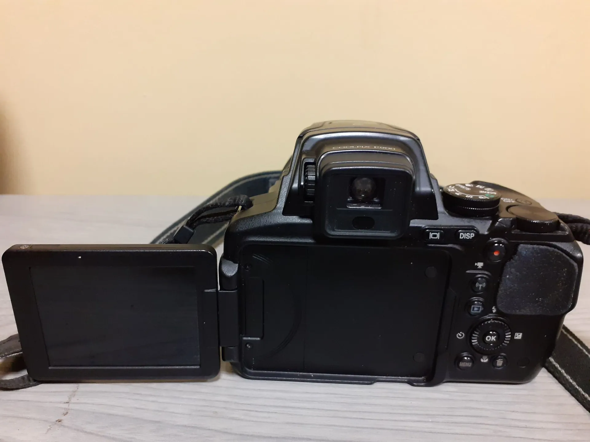 Czarny aparat fotograficzny marki Canon z wysuniętym do boku ekranem LCD, widoczny od tyłu. Na obudowie znajdują się przyciski sterujące, w tym przycisk OK oraz DISP. Aparat ma przymocowany czarny pasek na szyję i spoczywa na jasnej, drewnianej powierzchni na tle beżowej ściany.