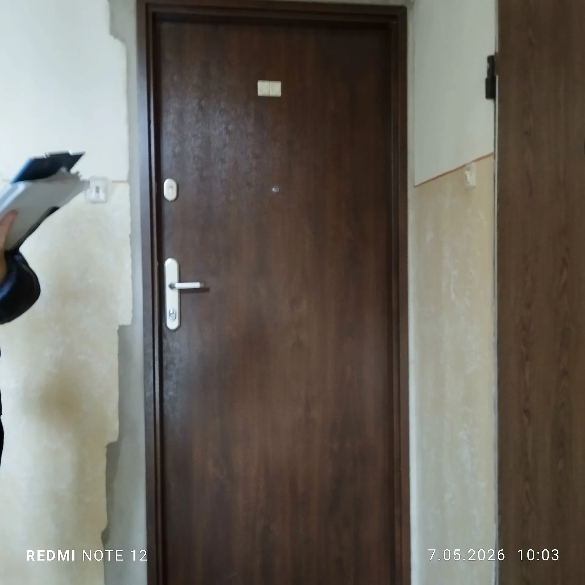 Zdjęcie przedstawiające ciemnobrązowe, drewniane drzwi wewnętrzne z klamką i otworem na klucz. Drzwi są osadzone w jasnej ścianie, która wygląda na nieukończoną lub w trakcie remontu, z widocznymi różnicami w kolorze tynku. Po lewej stronie widoczny jest fragment dłoni trzymającej biały przedmiot. W dolnej części obrazu znajduje się biały napis: 'REDMI NOTE 12' po lewej oraz data i godzina '7.05.2026 10:03' po prawej.