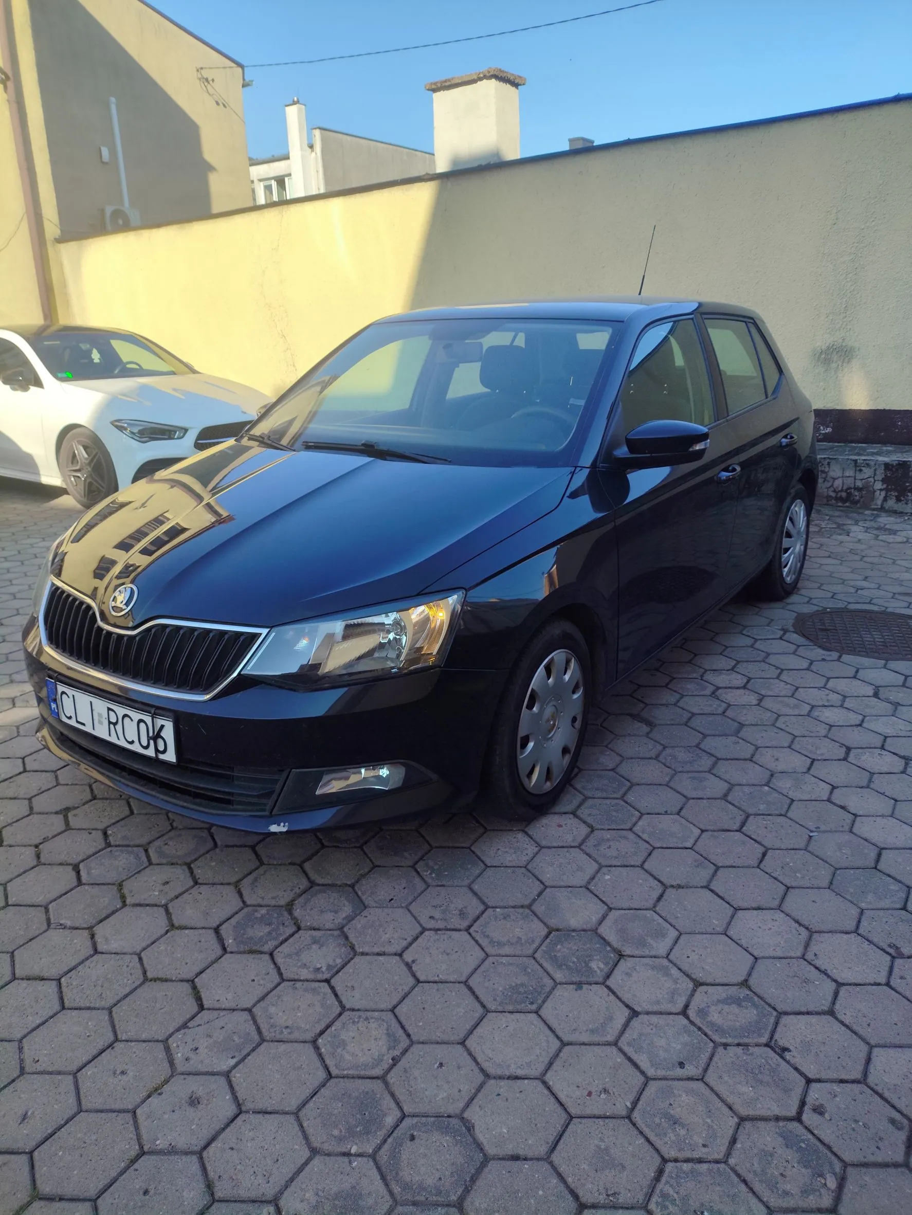 Czarny samochód marki Skoda zaparkowany na kostce brukowej o sześciokątnym kształcie. Auto jest widoczne z przodu i z boku. Na przednim zderzaku znajduje się tablica rejestracyjna o treści CLI RC06. W tle widoczny jest fragment białego samochodu oraz jasna ściana budynku pod błękitnym niebem.