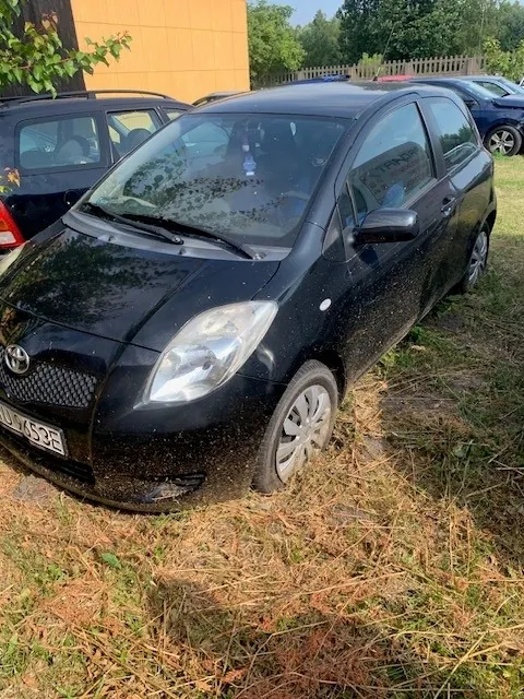 Czarny, trzy drzwiowy samochód osobowy marki Toyota zaparkowany na trawiastym podłożu z suchymi liśćmi. Widoczny jest przód i prawy bok auta. Samochód ma polską tablicę rejestracyjną o numerze kończącym się na 453E. W tle widoczny jest fragment żółtego budynku oraz inne zaparkowane samochody.