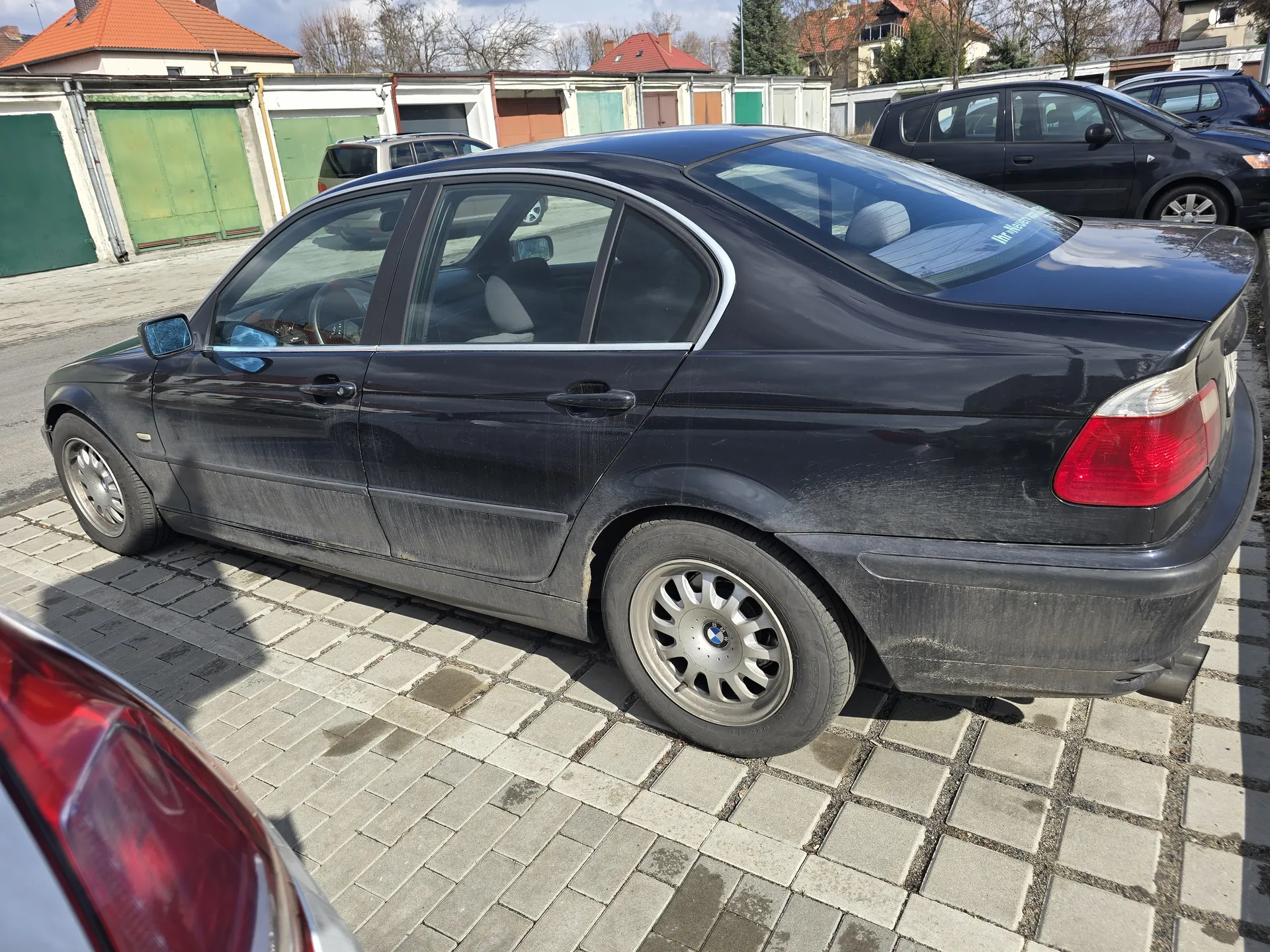 Czarny samochód osobowy marki BMW, czterodrzwiowy sedan, zaparkowany na brukowanej powierzchni. Pojazd jest zabrudzony kurzem. W tle widoczne garaże z kolorowymi bramami (zielone, czerwone, brązowe) oraz inne zaparkowane samochody. Na pierwszym planie fragment czerwonego światła tylnego innego pojazdu. Niebo częściowo zachmurzone.
