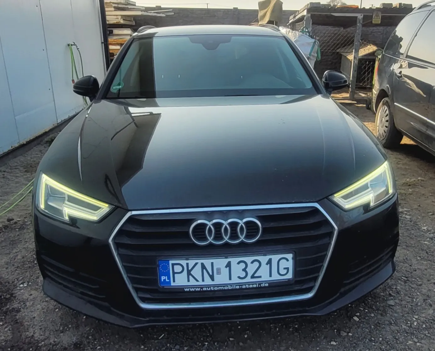 Czarny samochód marki Audi z przodu, z widoczną tablicą rejestracyjną PKN 1321G z niebieskim pasem PL po lewej stronie. Poniżej tablicy znajduje się napis www.automobile-atael.de. W tle widoczne są inne pojazdy, biały kontener, stosy materiałów i kamienna ściana. Samochód stoi na podłożu z żwiru, a reflektory są włączone.