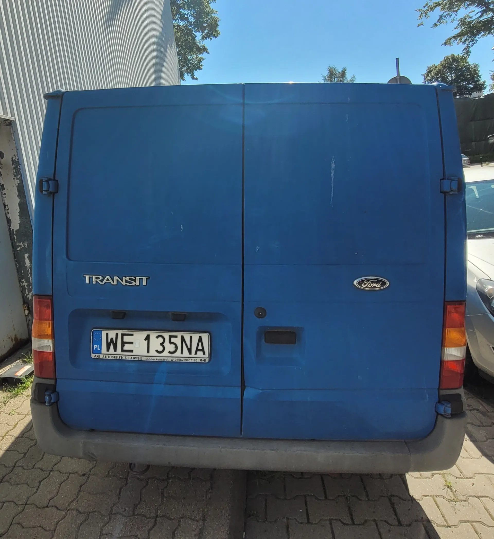 Tylny widok niebieskiego vana marki Ford Transit. Na lewych drzwiach znajduje się napis 'TRANSIT', a na prawych logo 'Ford'. Pojazd posiada polską tablicę rejestracyjną o numerze 'WE 135NA'. Van zaparkowany jest na kostce brukowej obok białego budynku.