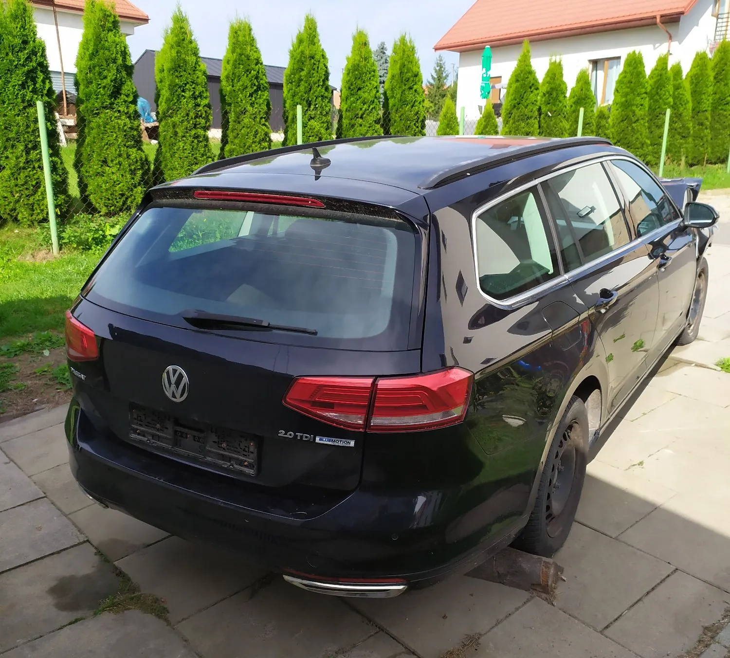Czarny samochód marki Volkswagen w nadwoziu kombi, zaparkowany na betonowym podjeździe. Widoczny jest tył i prawy bok pojazdu. Na klapie bagażnika znajduje się logo Volkswagena oraz oznaczenia '2.0 TDI' i '4MOTION'. W tle widać rządek wysokich, smukłych drzew iglastych oraz fragmenty białych domów z czerwonymi dachami.