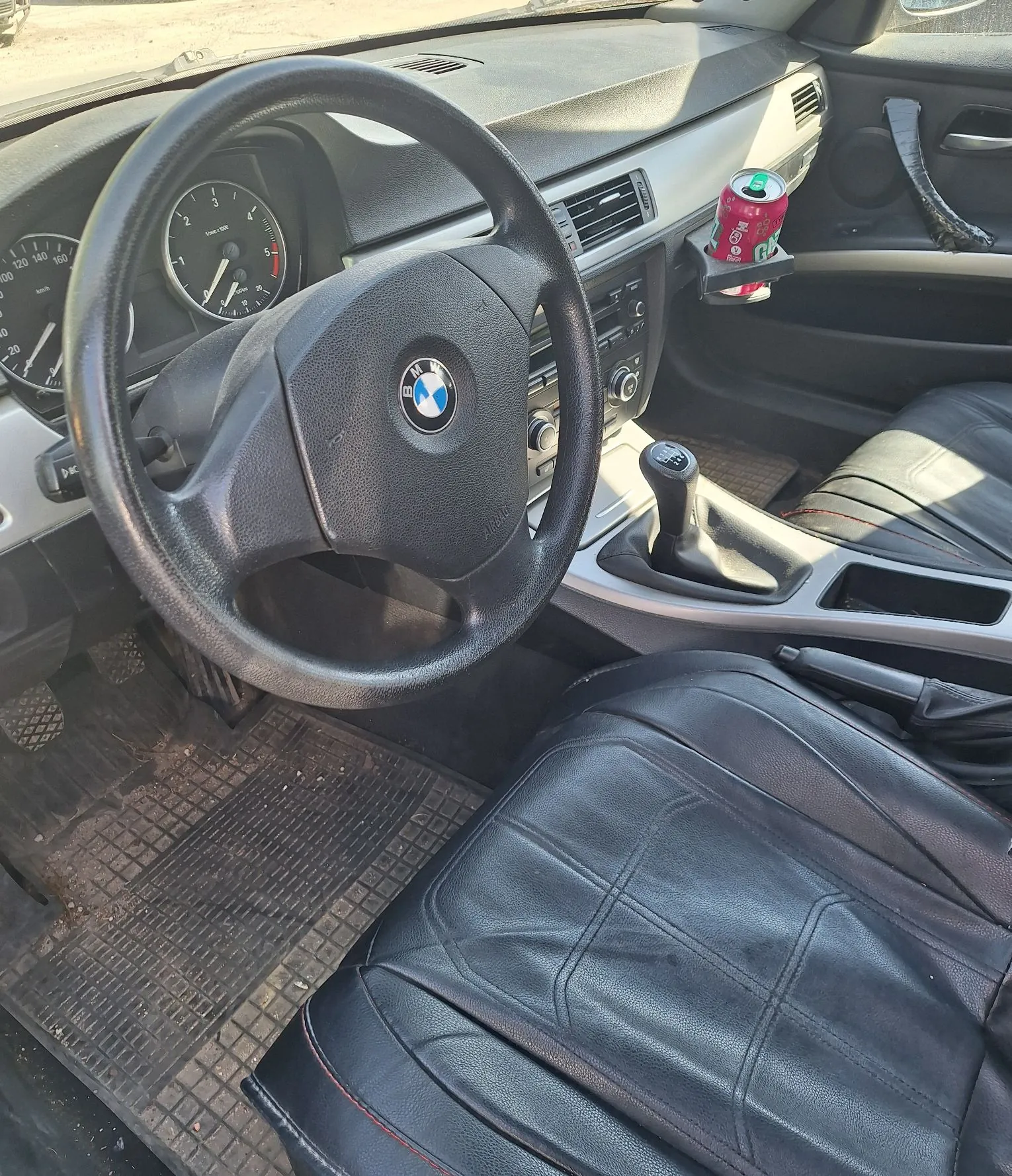 Wnętrze samochodu marki BMW widziane z perspektywy kierowcy. Widoczna jest czarna kierownica z logo BMW w centrum, deska rozdzielcza z zegarami, lewarek zmiany biegów oraz czarne, skórzane fotele z przeszyciami. W uchwycie przy konsoli środkowej znajduje się czerwona puszka napoju. Podłoga jest wyłożona gumowymi matami.