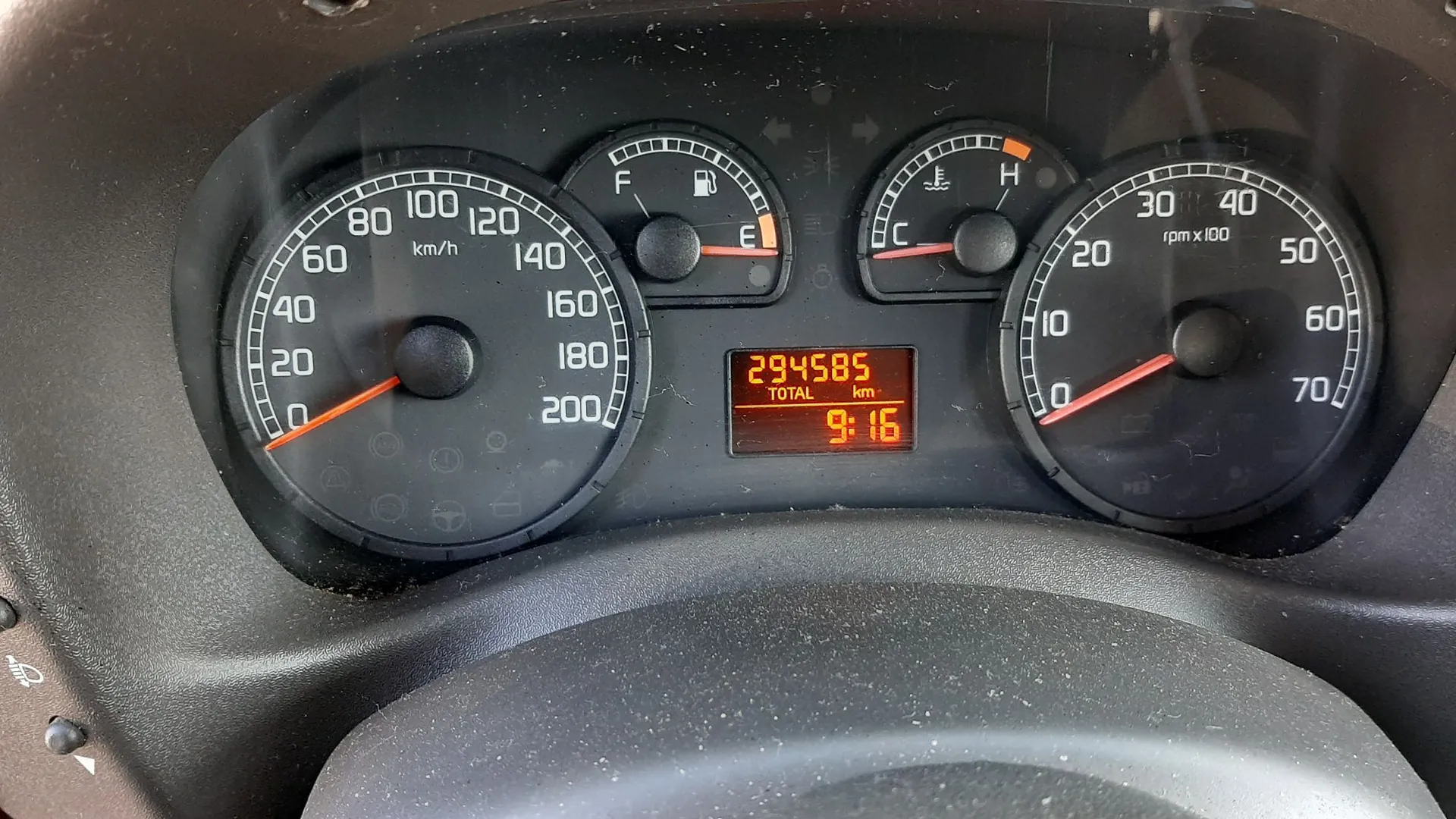 Zdjęcie deski rozdzielczej samochodu z analogowymi wskaźnikami. Po lewej stronie znajduje się prędkościomierz z zakresem do 200 km/h, w centrum wskaźnik poziomu paliwa (F-E) oraz temperatury silnika (C-H). Po prawej stronie znajduje się obrotomierz z zakresem do 70 x 100 rpm. W środkowej części widnieje cyfrowy wyświetlacz z pomarańczowymi cyframi wskazującymi całkowity przebieg 294585 km oraz godzinę 9:16.