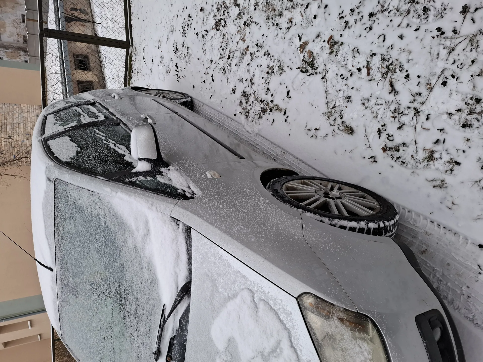 Srebrny samochód pokryty warstwą śniegu, zaparkowany na śnieżnej powierzchni z widocznymi plamami ziemi i suchych liści. W tle metalowe ogrodzenie siatkowe oraz budynki z ceglaną i beżową elewacją. Śnieg pokrywa przednią szybę, maskę, dach i boczne szyby pojazdu. Koła samochodu mają śnieg na oponach i felgach.