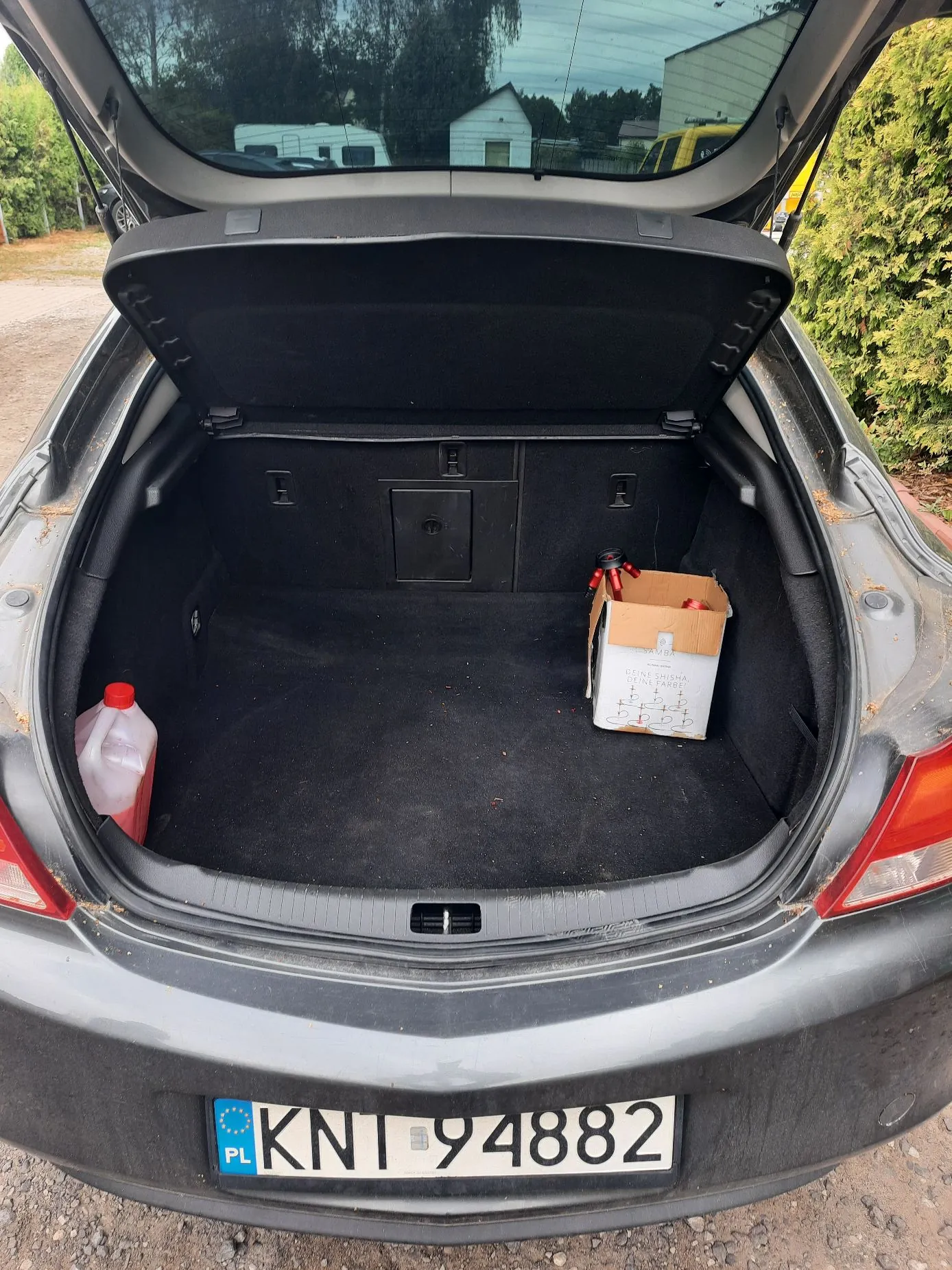 Widok z tyłu na otwarty bagażnik szarego samochodu. Wewnątrz bagażnika znajduje się biała plastikowa butelka z czerwoną zakrętką po lewej stronie oraz tekturowe pudełko z przedmiotami po prawej stronie. Na zderzaku samochodu widoczna jest polska tablica rejestracyjna o numerze KNI 94882.