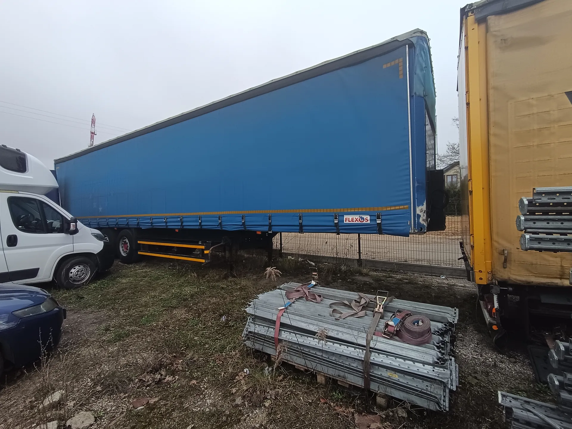 Na zdjęciu widoczne są pojazdy: niebieska naczepa z napisem FLEXOS, biała furgonetka, fragment ciemnoniebieskiego samochodu i żółta naczepa. W pierwszym planie leżą metalowe konstrukcje związane paskami. Tło stanowi ogrodzenie, trawa, drobne kamienie i budynki. Niebo jest pochmurne.