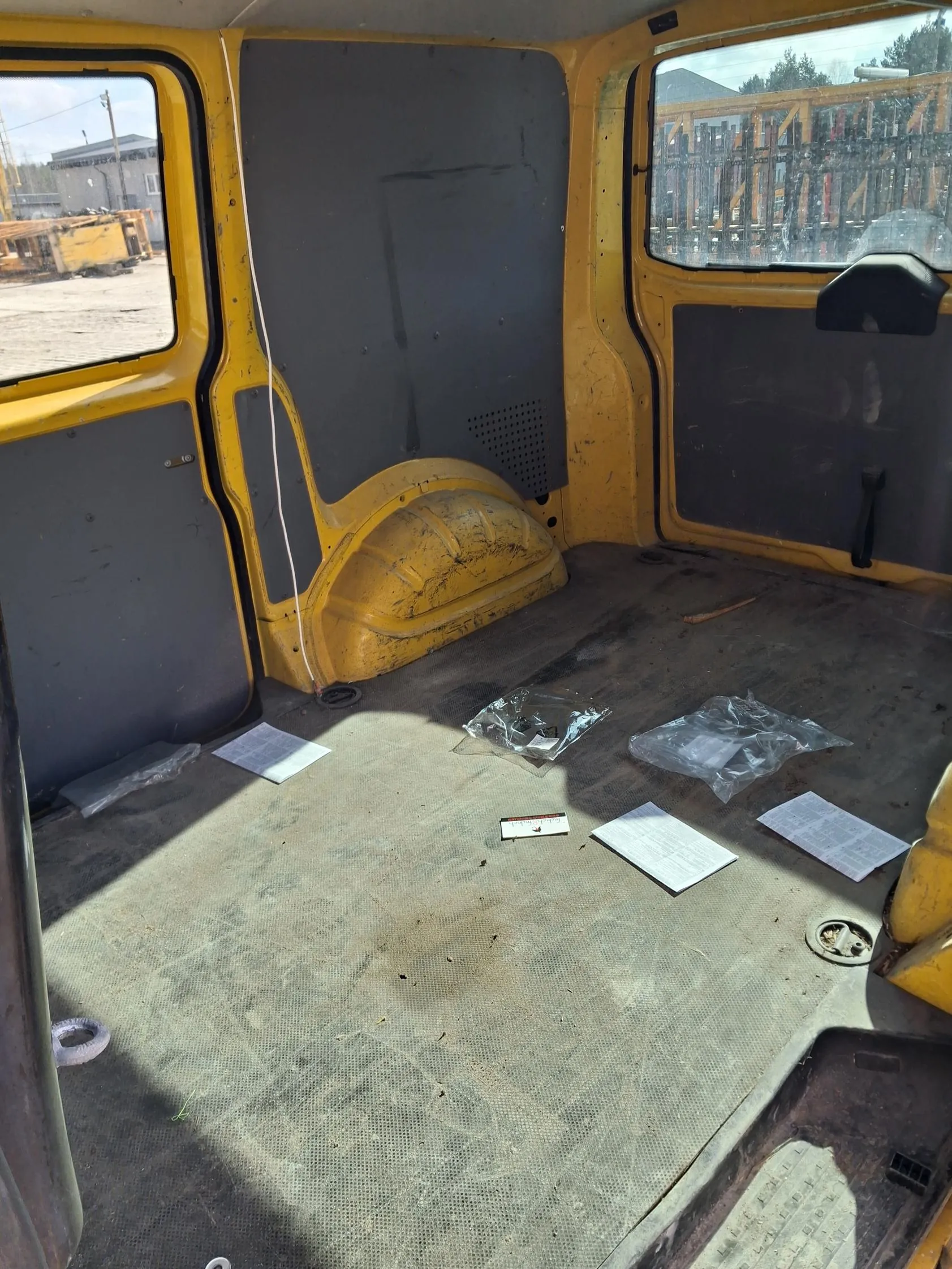 Wnętrze żółtego pojazdu z ciemnoszarymi panelami. Widoczne okna po obu stronach, przez które widać teren budowlany z maszynami i budynkami. Na podłodze leżą kilka kartek papieru, przezroczyste torebki i mała karta. Podłoga jest zabrudzona, a ściany mają ślady zużycia.