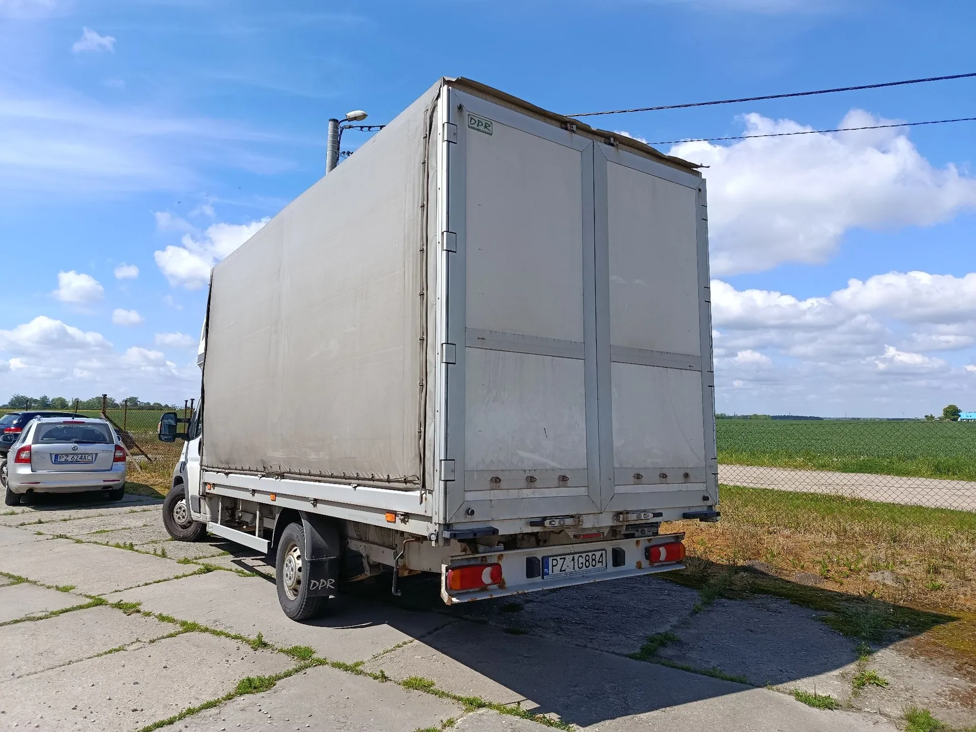 Biała ciężarówka z zabudową furgonową zaparkowana na betonowym placu. Na tylnych drzwiach pojazdu widoczna jest tablica rejestracyjna o numerze PZ 1G884. W tle po lewej stronie stoi srebrny samochód osobowy z tablicą rejestracyjną PZ 824CGE. W oddali widać zielone pole pod niebem z białymi chmurami.