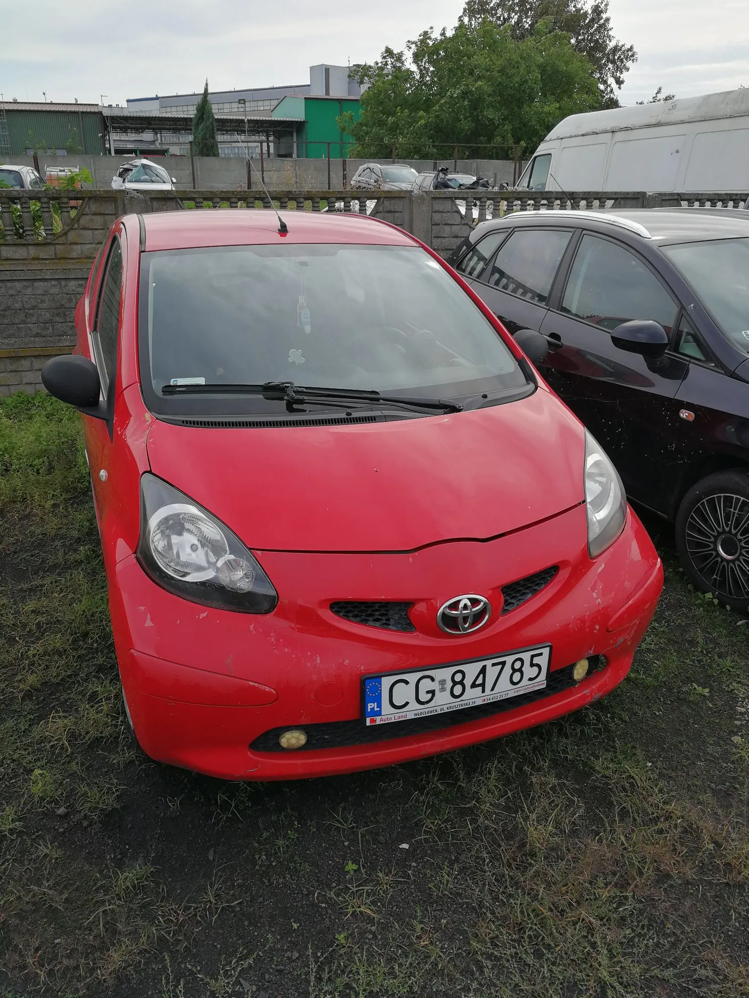Czerwony samochód osobowy marki Toyota zaparkowany na trawiastym i piaszczystym podłożu. Pojazd ma widoczną tablicę rejestracyjną o numerze CG 84785 z oznaczeniem PL. Obok stoi ciemny samochód, a w tle widać betonowy płot, inne zaparkowane auta oraz budynki przemysłowe i drzewa pod zachmurzonym niebem.