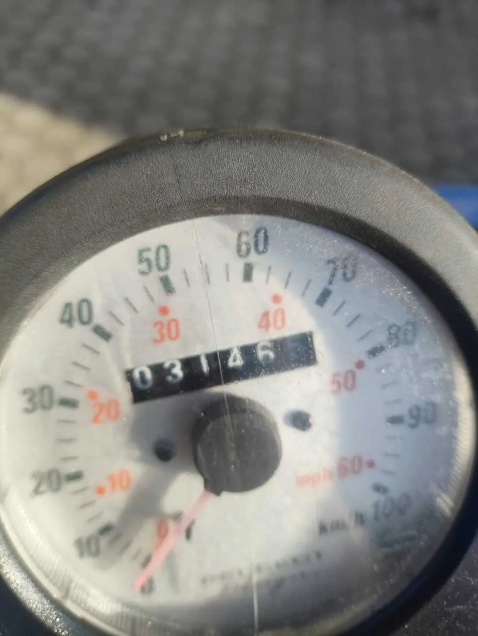 Zbliżenie na licznik prędkości z cyfrowym przebiegiem 03146. Skala licznika obejmuje wartości od 0 do 100, z oznaczeniami w km/h i mph. Widoczne są liczby 10, 20, 30, 40, 50, 60, 70, 80, 90, 100 oraz czerwone punkty przy 30, 40, 50. Wskazówka znajduje się blisko zera. Tło jest rozmyte, przypominające nawierzchnię asfaltową.