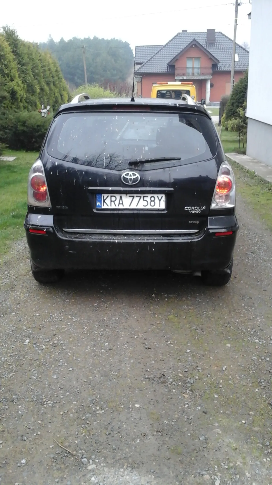 Z tyłu czarnego samochodu marki Toyota Corolla widoczna tablica rejestracyjna KRA 7758Y. Samochód zaparkowany na żwirowej powierzchni. W tle czerwony dom z ciemnym dachem, zieleń oraz żółty pojazd. Na szybie i nadwoziu samochodu widoczne krople wody.