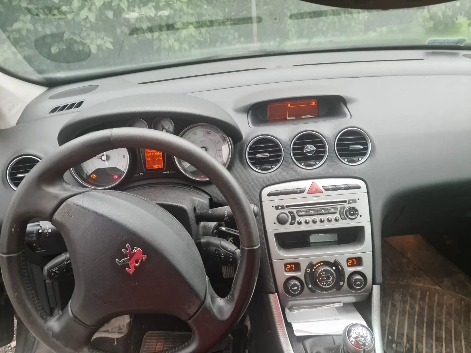 Wnętrze samochodu z widocznym kierownicą z czerwonym logo lwa, tablicą przyrządów z analogowymi wskaźnikami prędkości i obrotomierza, cyfrowym wyświetlaczem z pomarańczowym podświetleniem, trzema okrągłymi wydmuchami powietrza, centralnym panelem z radiem, przyciskami sterującymi klimatyzacją oraz pokazującym temperaturę 27. W tle przez przednią szybę widoczne są zielone liście drzew. Wnętrze jest w kolorze szarym, a na desce rozdzielczej znajduje się wyświetlacz z pomarańczowym oświetleniem.