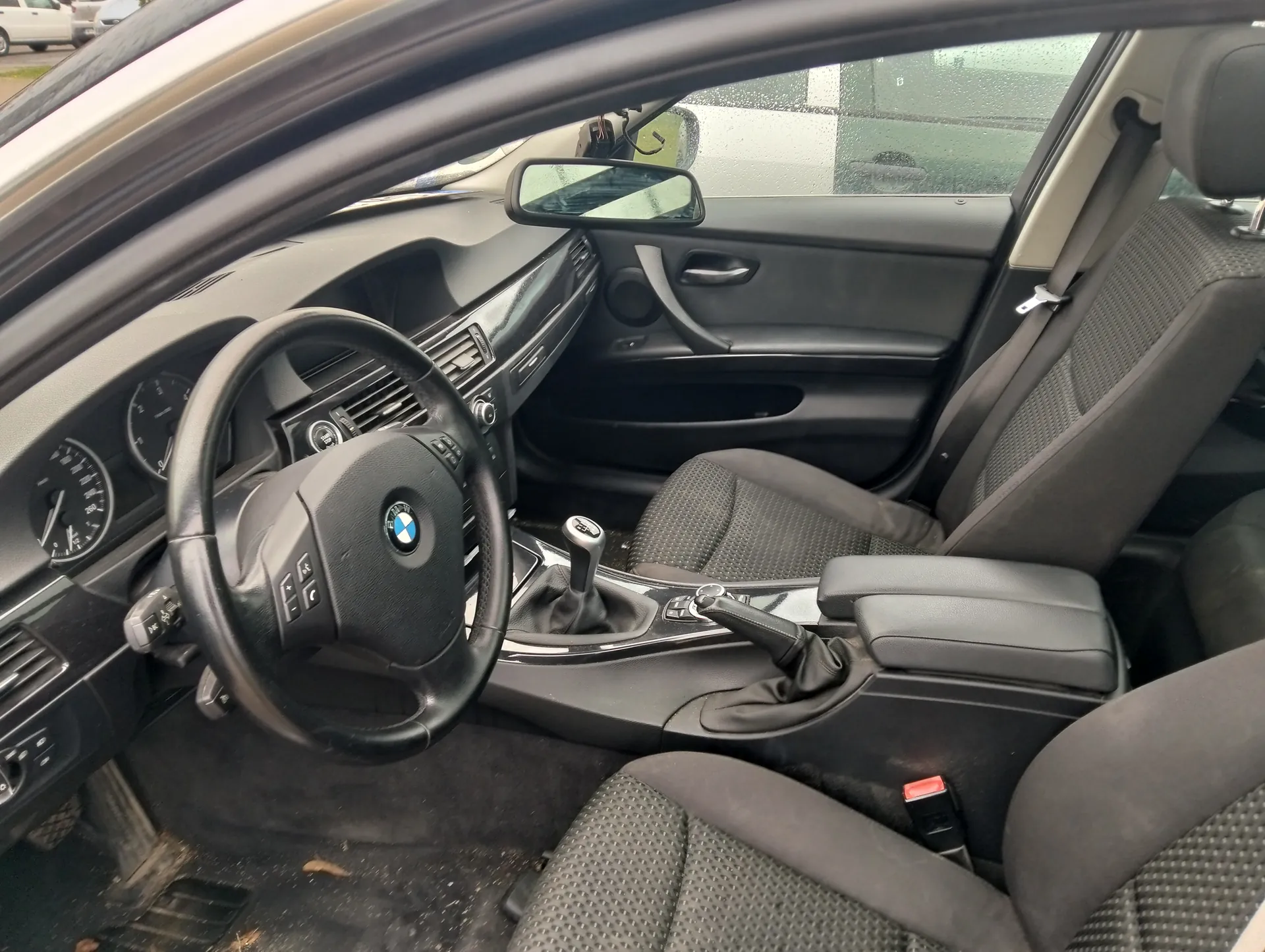 Wnętrze samochodu marki BMW z widocznym kierownicą z logo BMW, czarnymi siedzeniami z perforowaną tapicerką, deską rozdzielczą z licznikami, dźwignią zmiany biegów i centralną konsolą. Widoczne są również lusterko boczne, drzwi pasażera z uchwytem oraz fragment zewnętrzny przez przednią szybę z innym pojazdem i budynkiem w tle.