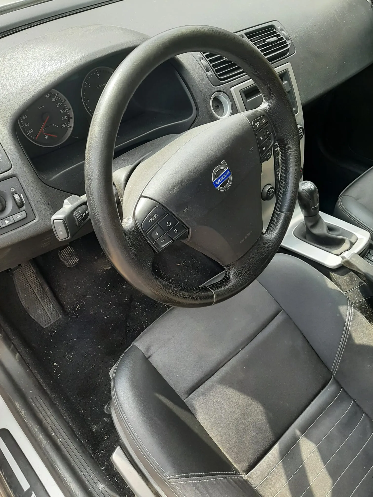 Wnętrze samochodu Volvo z widocznym kierownicą z logo marki, przyciskami sterowania na kierownicy, zegarami przyrządów (w tym prędkościomierzem do 260 km/h i tachometrem), czarnymi skórzonymi fotelami, dźwignią zmiany biegów oraz czarną podłogą z widocznymi śladami brudu. Deska rozdzielcza i inne elementy są w odcieniach szarości i czerni.