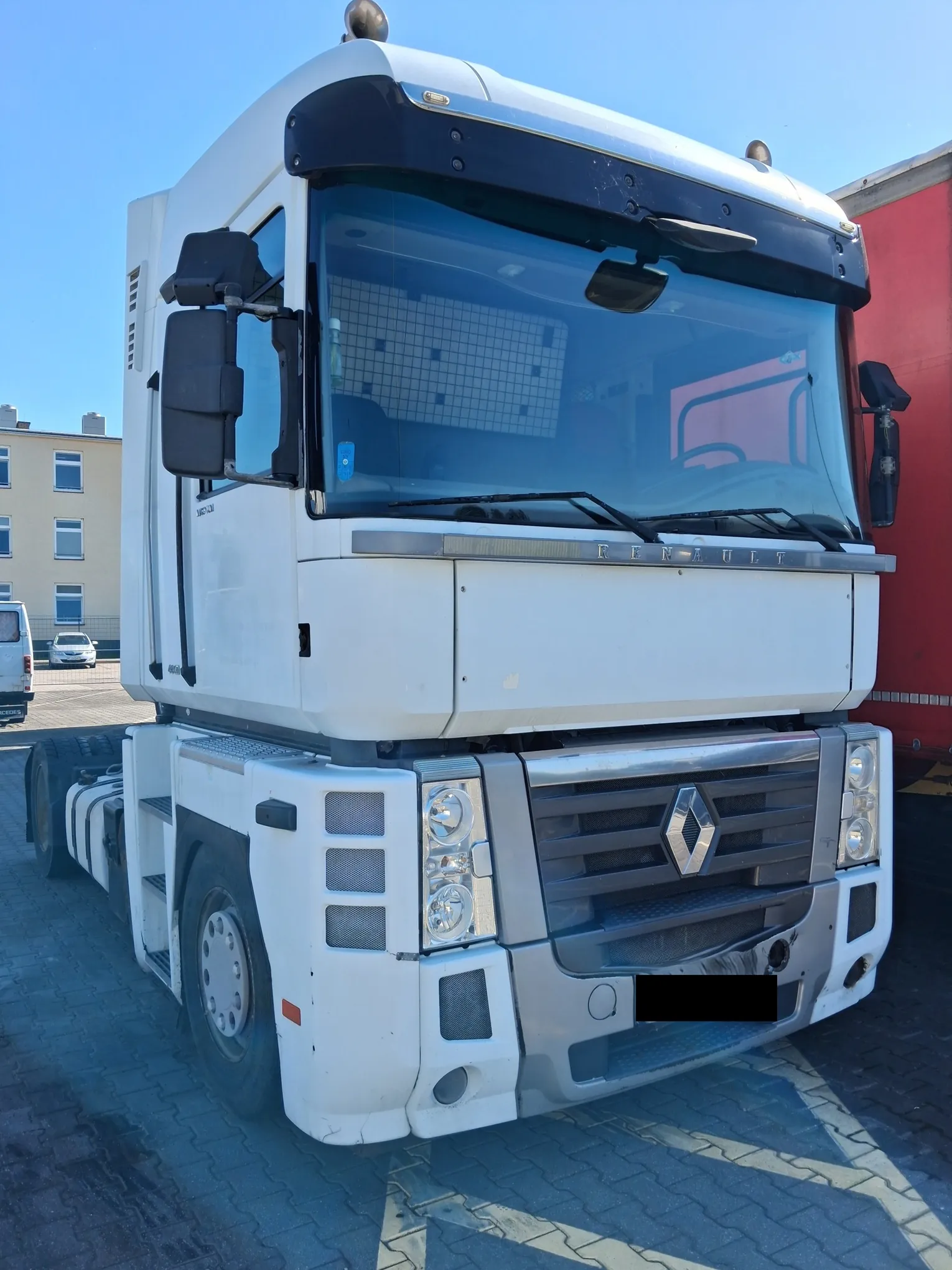 Biała ciągnik siodłowy marki Renault widziany z przodu i z lewej strony. Pojazd posiada dużą przednią szybę, charakterystyczny grill z logo Renault w centrum oraz zestaw reflektorów. Ciągnik stoi na kostce brukowej na tle jasnego budynku i niebieskiego nieba.
