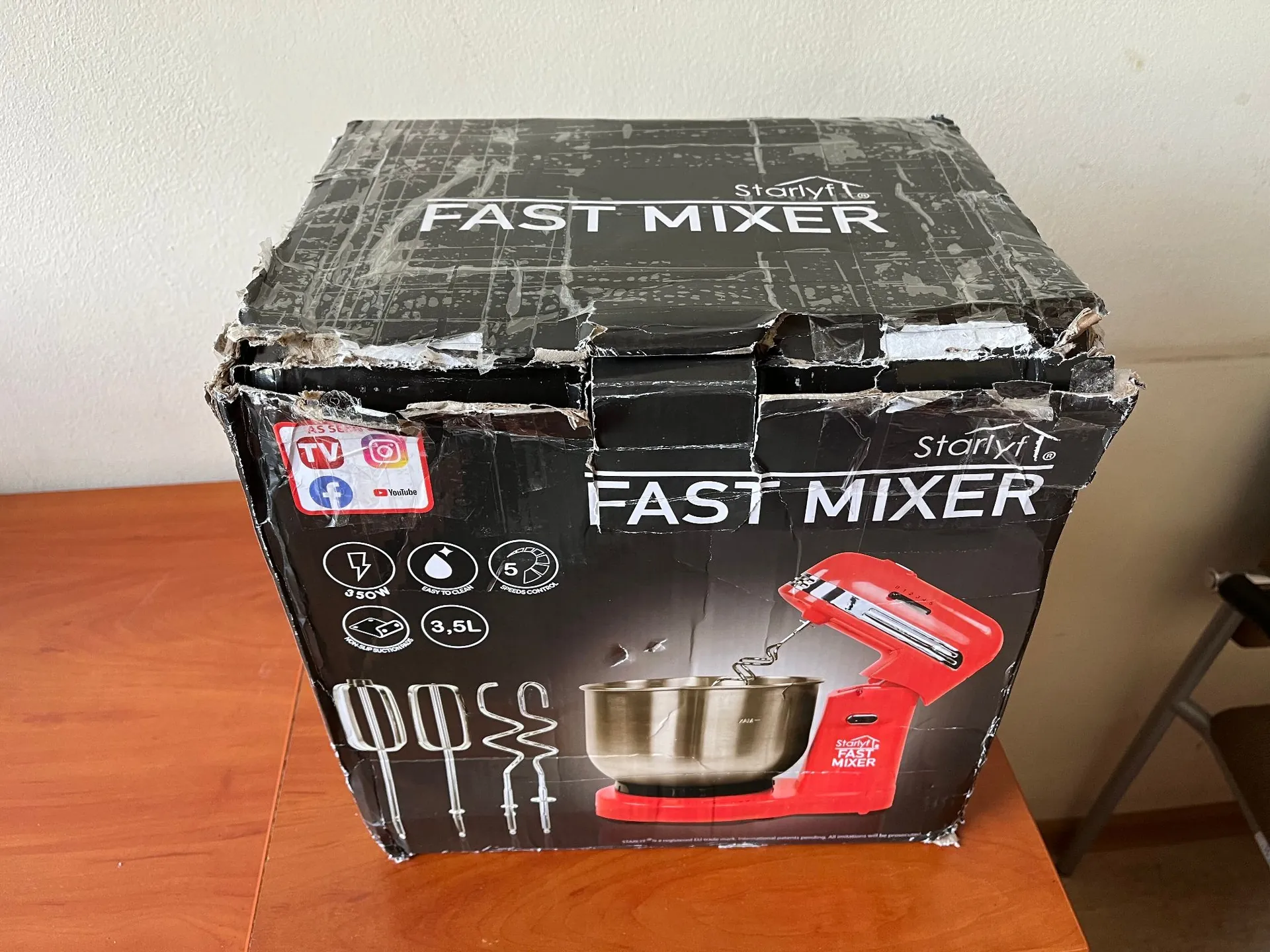 Zdjęcie uszkodzonego, czarnego kartonowego opakowania miksera stacjonarnego, stojącego na drewnianym blacie. Na pudełku widnieje biały napis „FAST MIXER” oraz logo marki „Starlyf”. Na przedniej ściance znajduje się zdjęcie czerwonego miksera z metalową misą oraz zestawem końcówek mieszających. Widoczne są również ikony funkcji: 350W, „easy to clean”, „speed control” oraz pojemność „3,5L”. Górna część pudełka jest mocno pognieciona i przerwana.