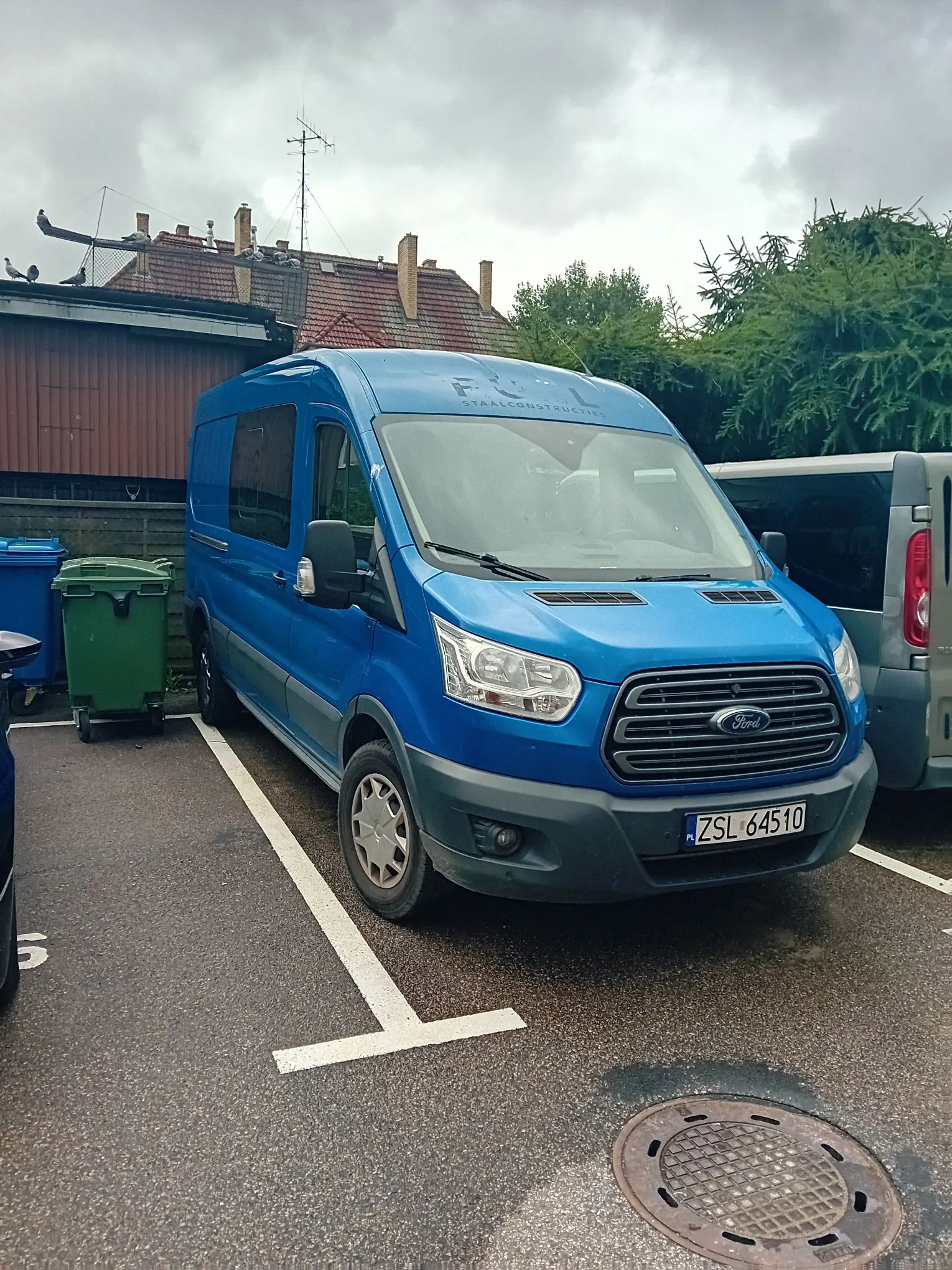 Niebieski furgon marki Ford zaparkowany na asfaltowym parkingu. Na przedniej części dachu pojazdu widnieje napis 'F U L STAALCONSTRUCTIES'. Samochód ma tablicę rejestracyjną o numerze ZSL 64510. W tle widoczne są zielone kosze na śmieci, budynek z brązowym dachem oraz drzewa pod zachmurzonym niebem.