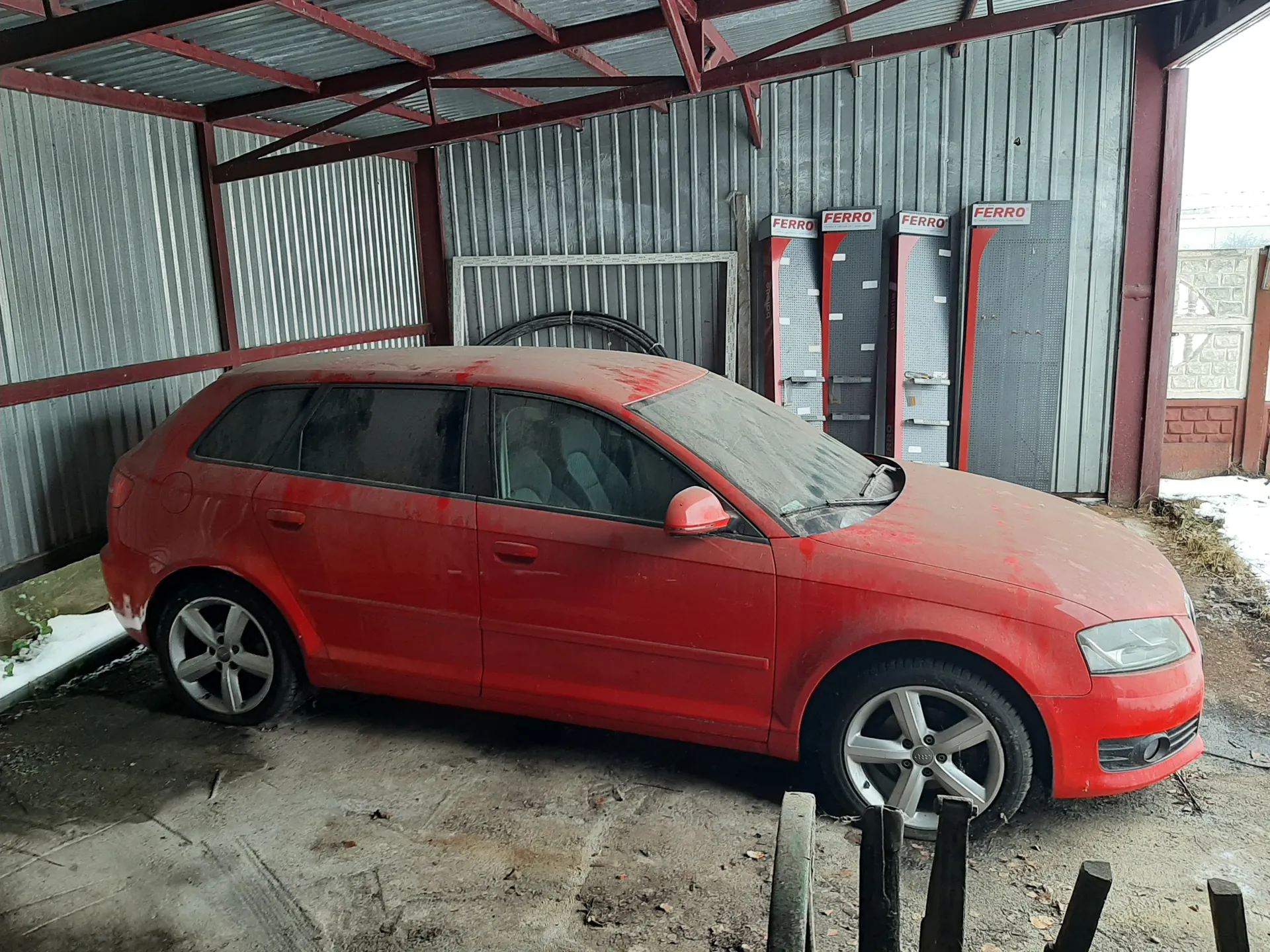 Czerwony samochód osobowy typu kombi zaparkowany pod metalowym daszkiem. Samochód pokryty jest kurzem, widoczne są srebrne felgi i ciemne szyby. W tle metalowa ściana z czterema czerwono-szarymi panelami z napisem FERRO. Na ziemi betonowej widoczne są śnieg, sucha trawa i fragmenty opony opartej o ścianę. W dolnej części kadru znajdują się czarne słupki ogrodzenia.