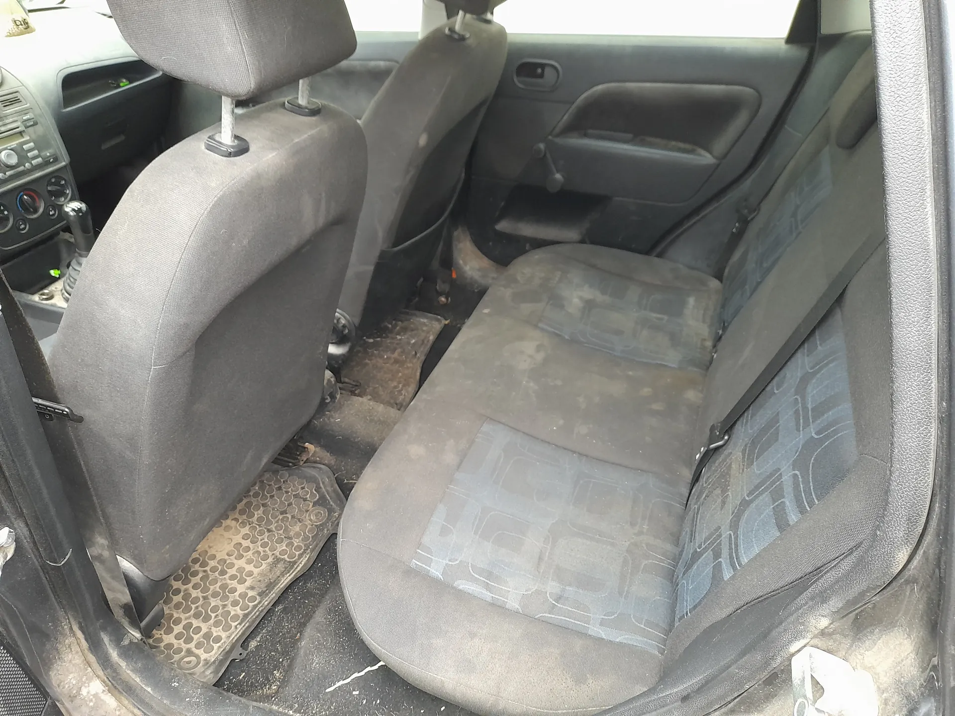 Wnętrze samochodu z widocznymi przodem siedzeniami pasażerskimi i kierowcy. Siedzenia są szare, z niebieskimi geometrycznymi wzorami, zabrudzone i zużyte. Widoczna jest deska rozdzielcza z przyciskami, dźwignia zmiany biegów po lewej stronie oraz brudne dywaniki podłogowe. Drzwi pasażerskie z uchwytem i okienkiem są po prawej stronie kadru. Całość prezentuje się w stanie nieczystym, z widocznymi plamami i zabrudzeniami na siedzeniach i podłodze.