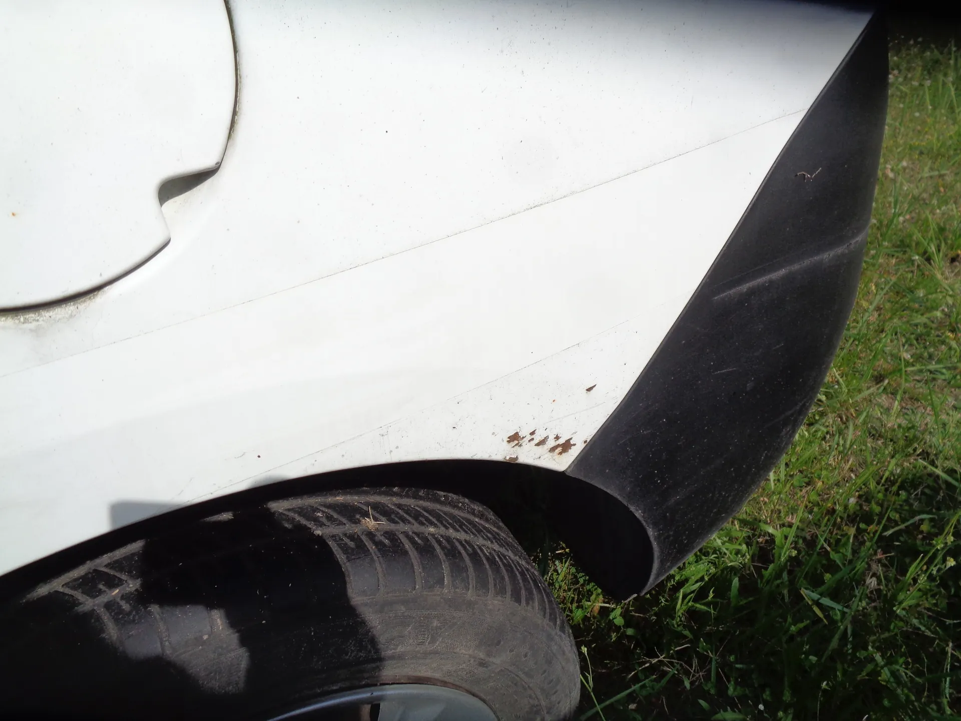 Zbliżenie na tylny błotnik białego samochodu. Widoczna jest czarna plastikowa osłona zderzaka oraz fragment opony. Na białym lakierze, tuż nad nadkolem, znajdują się niewielkie ślady rdzy i odpryski.