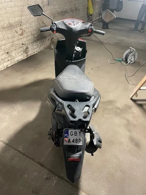 Czarny skuter widziany od tyłu z tablicą rejestracyjną PL GBY A48P. Skuter stoi na betonowej podłodze w garażu z ceglaną ścianą po lewej stronie. W tle widoczne są przewody, zwinięty kabel oraz osoba stojąca blisko drzwi. Lusterka boczne i siedzenie skutera są wyraźnie widoczne.