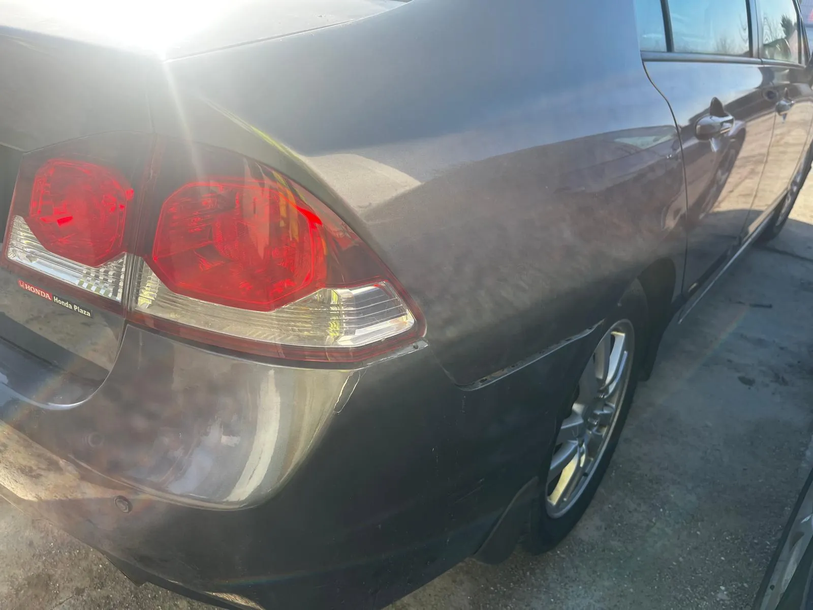 Zbliżenie na tylną część czarnego samochodu marki Honda. Widoczne są czerwone światła tylnego oświetlenia, chromowana obudowa reflektorów oraz napisy 'HONDA' i 'Honda Plaza' na lewej stronie bagażnika. Samochód stoi na betonowej powierzchni, a w tle widać odbicia słońca na karoserii.