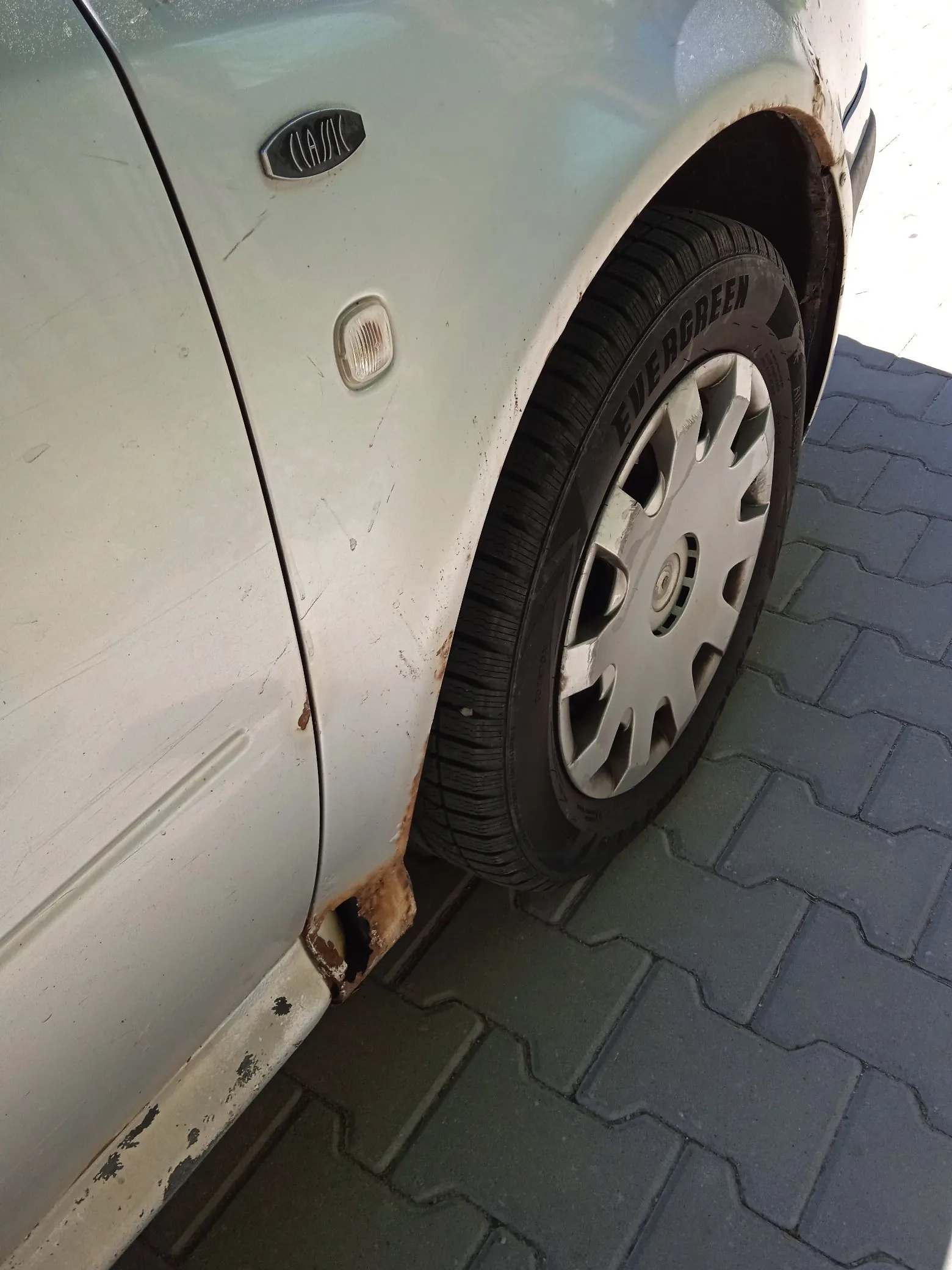 Zbliżenie na przedni lewy błotnik i koło jasnoniebieskiego samochodu. Na nadkolu oraz w dolnej części błotnika widoczne są znaczne wżery i rdza. Opona ma napis 'EVERGREEN' i jest zamontowana na srebrnej feldze z kołpakiem. Samochód stoi na szarej kostce brukowej. Na błotniku znajduje się mały, owalny emblemat z napisem 'CLASSIC' oraz boczny kierunkowskaz.
