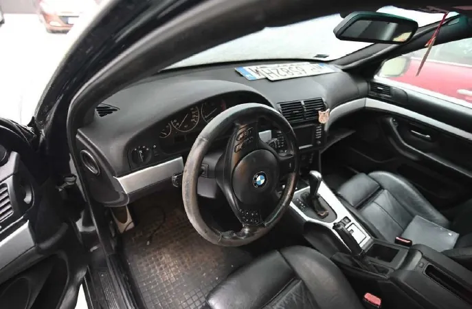 Wnętrze samochodu marki BMW z widocznym kierownicą z logo, czarnymi skórzonymi fotelami, deska rozdzielcza z licznikami i tablicą rejestracyjną K47857 umieszczoną na desce. Drzwi po stronie kierowcy są otwarte, widoczny jest pedał gazu i hamulca oraz centralna konsola z dźwignią zmiany biegów. Poza samochodem widoczne są fragmenty innych pojazdów.