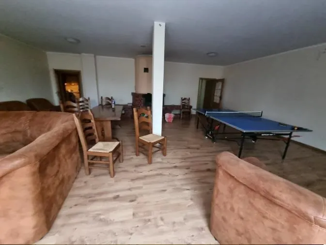 Wielka sala z brązowymi sofami po lewej i prawej stronie, drewnianymi krzesłami i stołem w centrum, niebieskim stołem do tenisa stołowego po prawej stronie, białą kolumną w środku pomieszczenia, podłogą z drewnianych desek oraz drzwiami w tle. Ściany są jasne, a sufit posiada okrągłe oświetlenie.